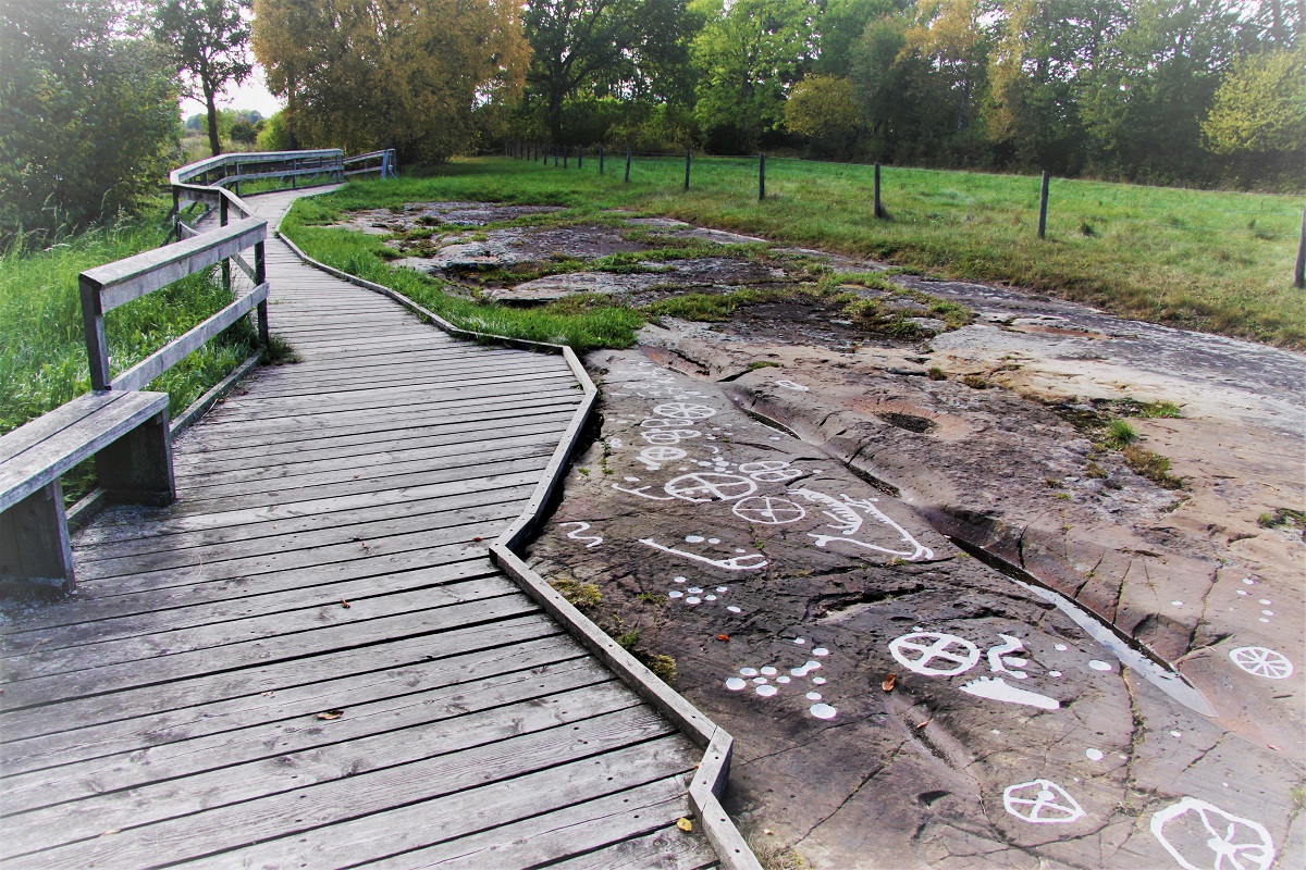 Gång av brädor som sträcker sig till vänster i bilden, med en bänk i nedre hörnet. Till höger finns klipphällar med hällristningar på.