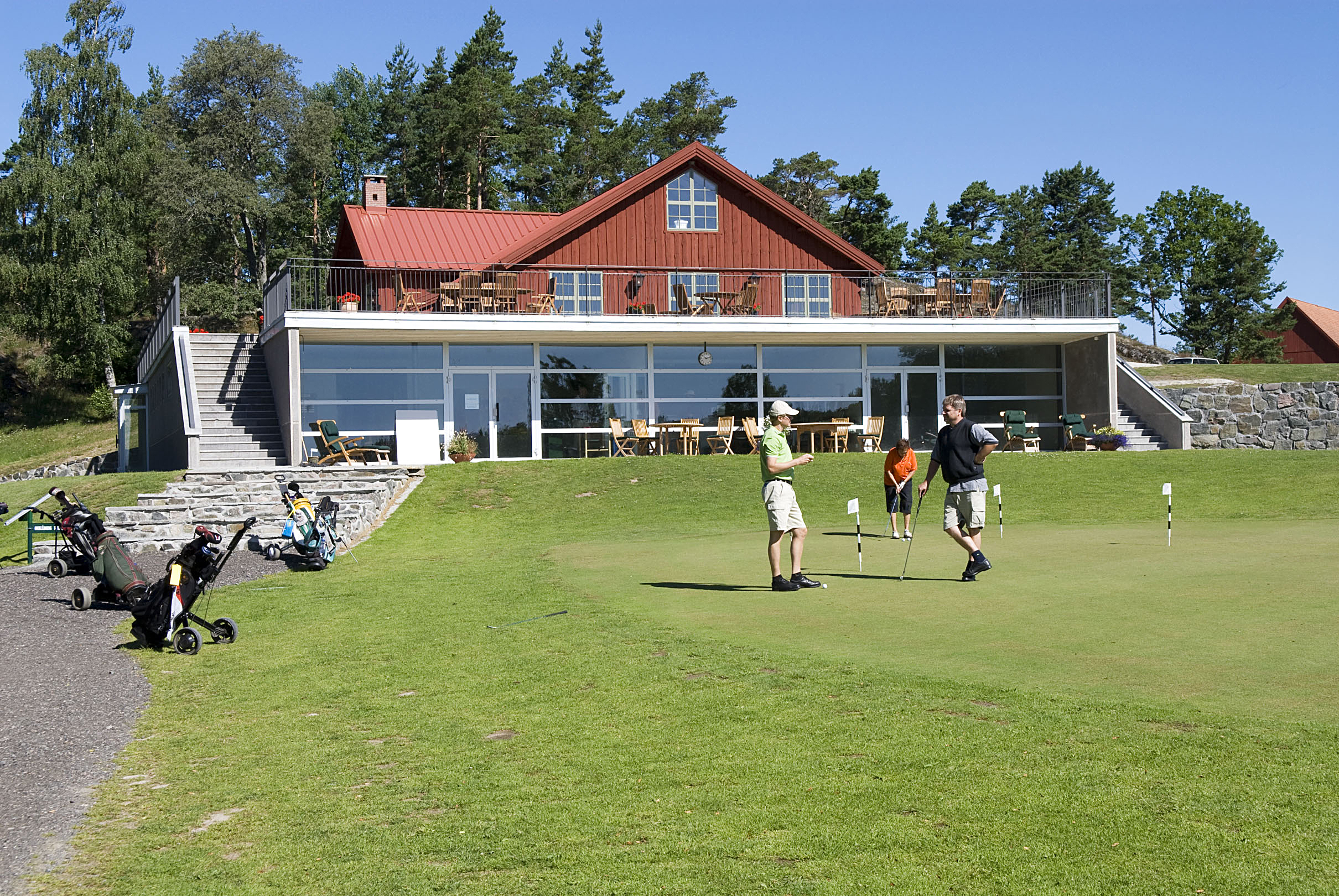 En golfanläggning och utanför står några personer och pratar med varandra redo att starta sin golfrunda på greenen.