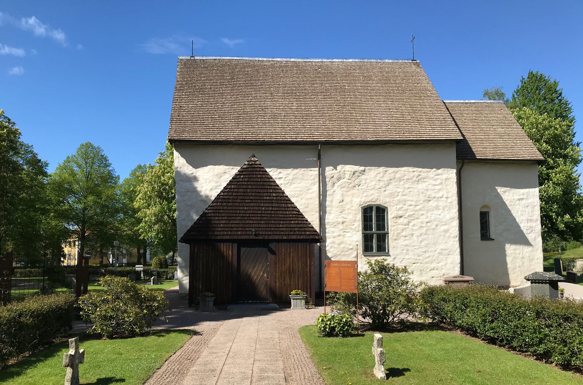 Götene kyrka utvändigt. Vit stenbyggnad med brunt trähus som ingång. En gång av stenplattor leder fram till kyrkan. 