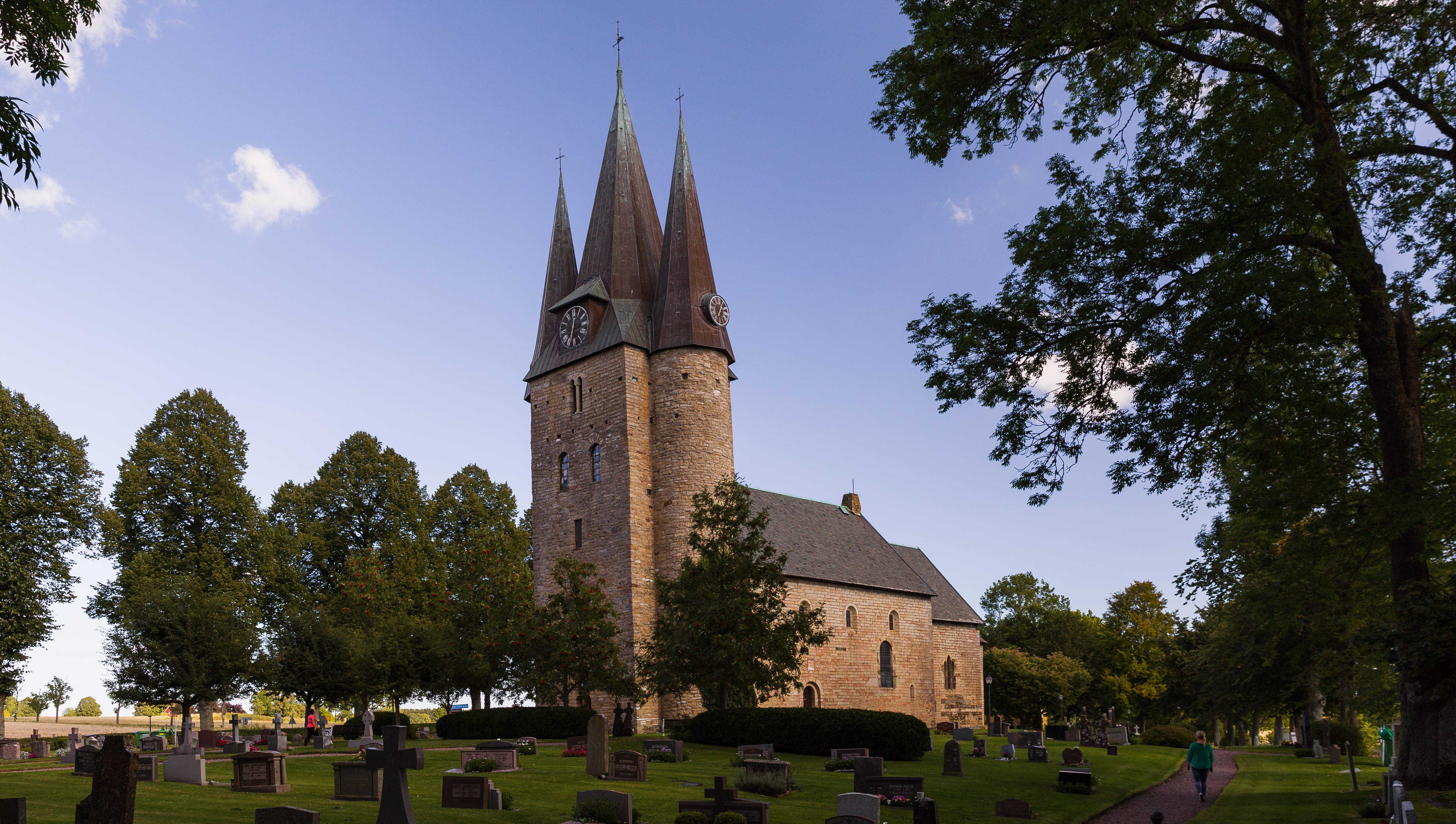 Gammal kyrka med tre torn. Framför kyrkan är det en kyrkogård med gravstenar och träd.