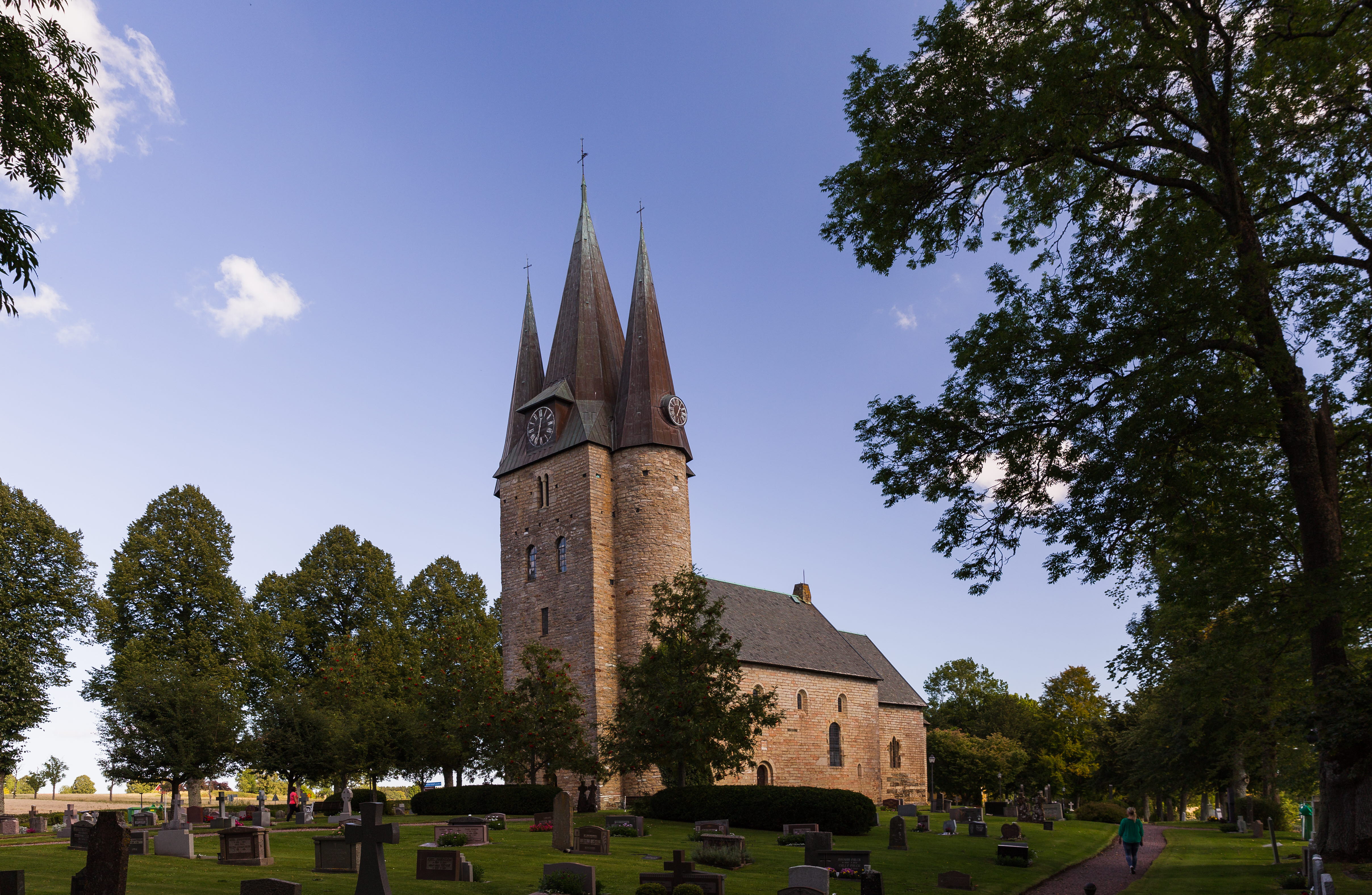 Gammal kyrka med tre torn. Framför kyrkan är det en kyrkogård med gravstenar och träd.