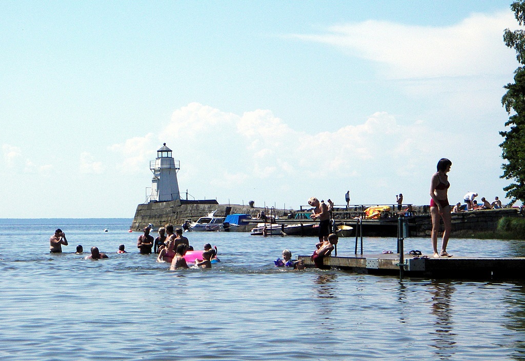 Flera människor som badar från en badbrygga. I bakgrunden syns en fyr på en pir. Det är en vacker sommardag.