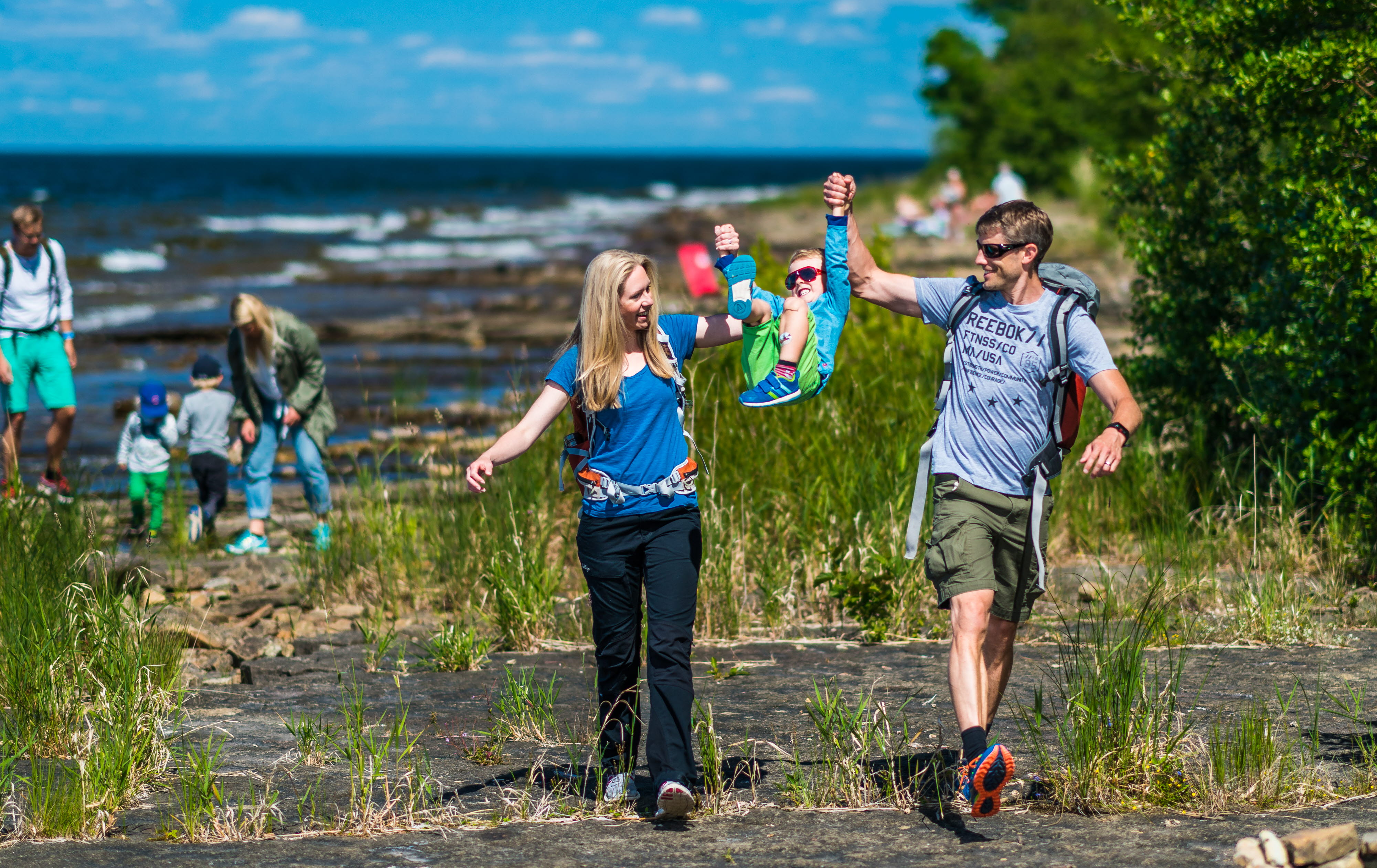En mamma och en pappa lyfter upp sin son i händerna i framtraskande på klipporna längs med strandkanten. 