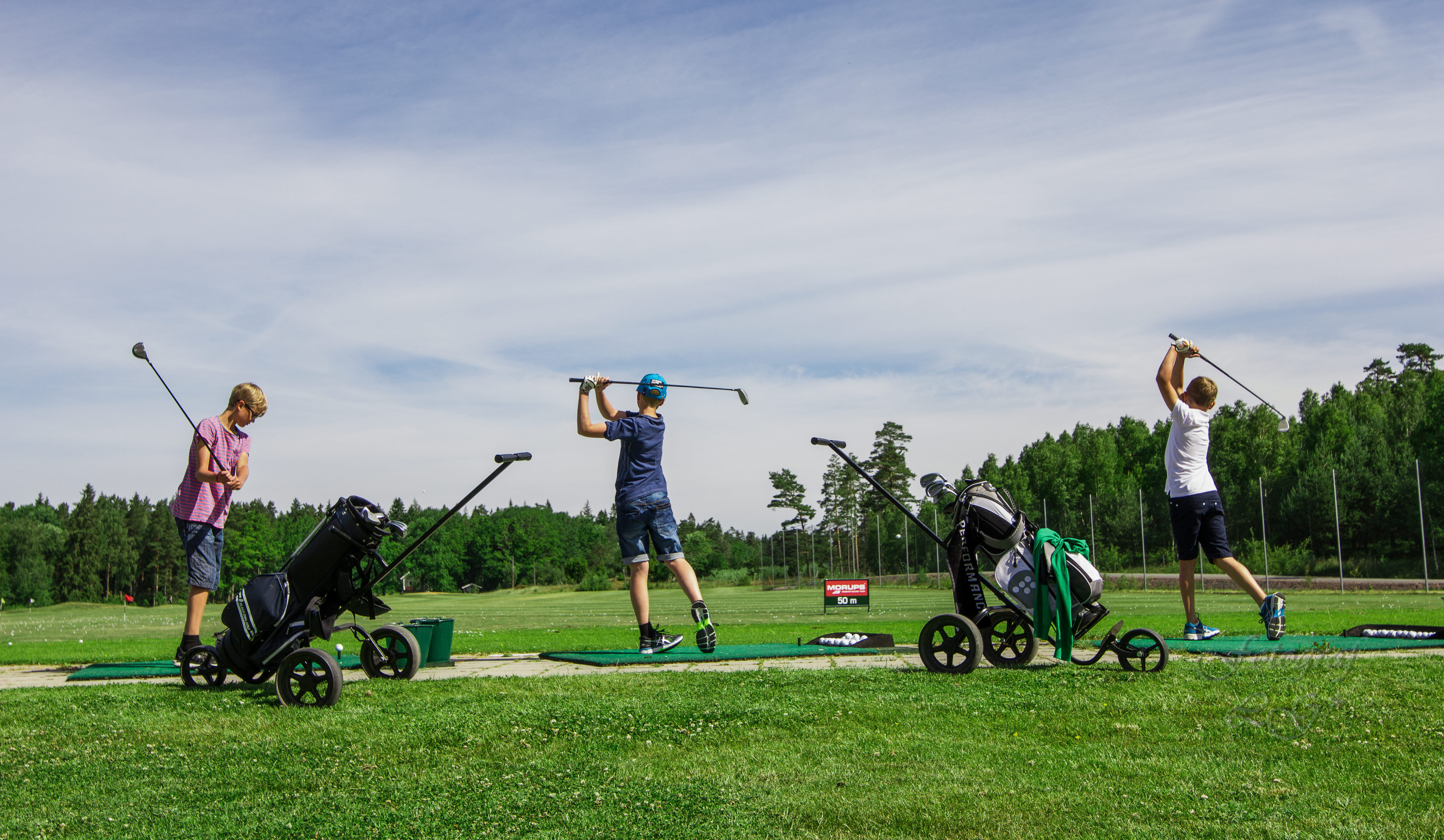 Some children stand and hit a golf course on a sunny summer day.