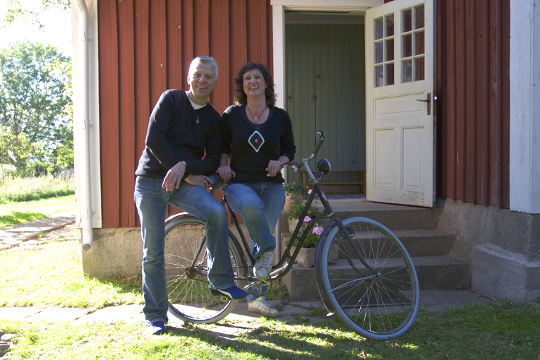 En man och en kvinna står vid en cykel och ler. I bakgrunden finns en förstukvist med en trappa och en vit dörr som är öppen till ett hus som är rött med vita knutar.