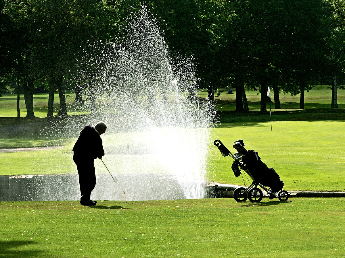 En man står på en green och golfar. I bakgrunden syns en fontän spruta och bredvid honom står hans golfvagn.