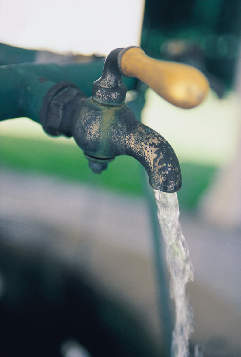 Old tap with water flowing out. Green pipes in the background and a yellow handle above the tap.