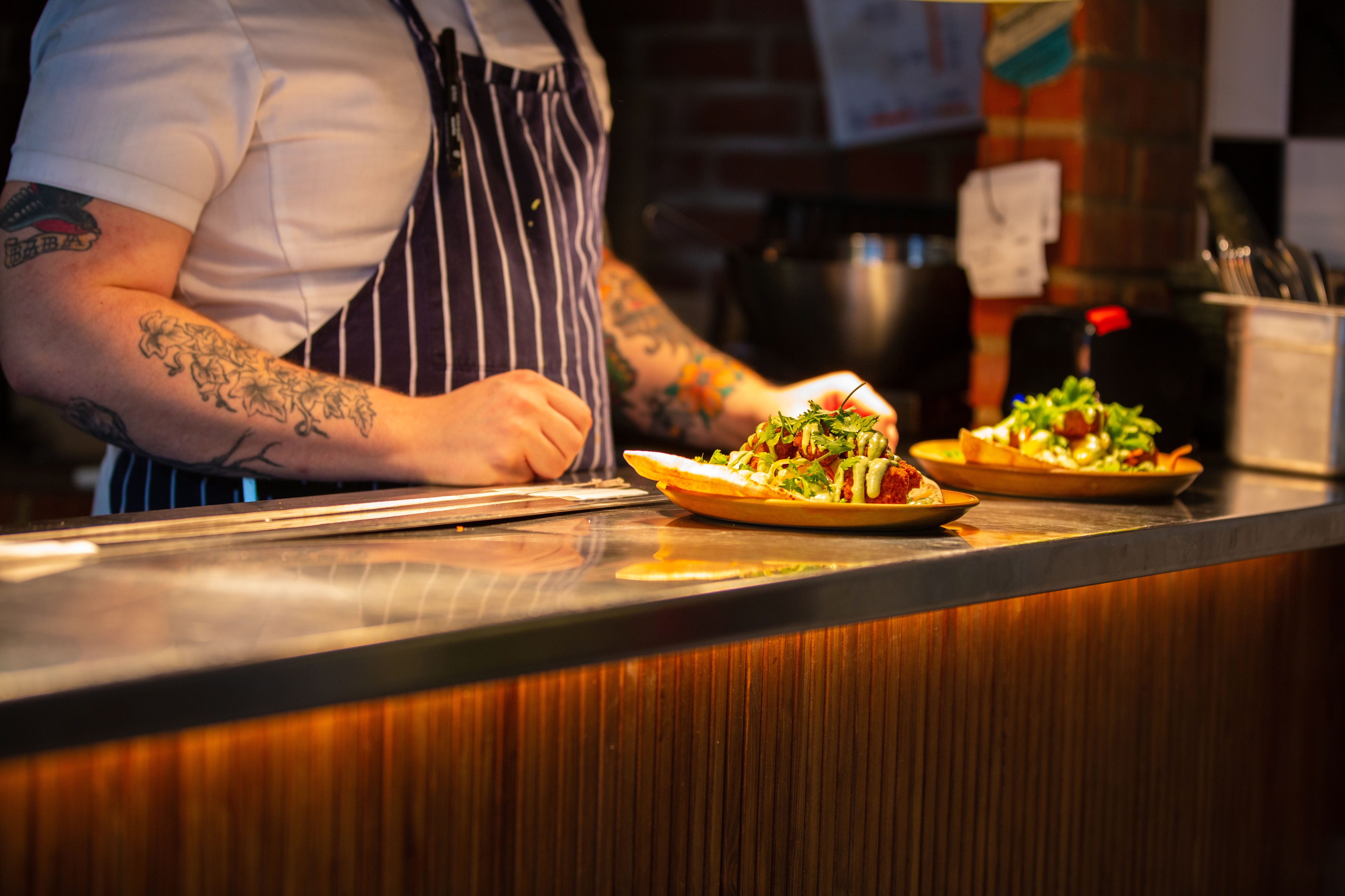 En manlig kock med tatuering på armarna står vid en bänk med mat upplagd på tallrikar.