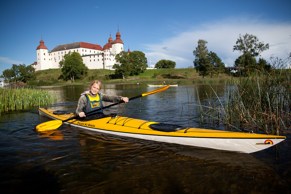 En kvinna sitter i en kajak och tittar in i kameran. Bakom henne syns pampiga vita Läckö Slott.