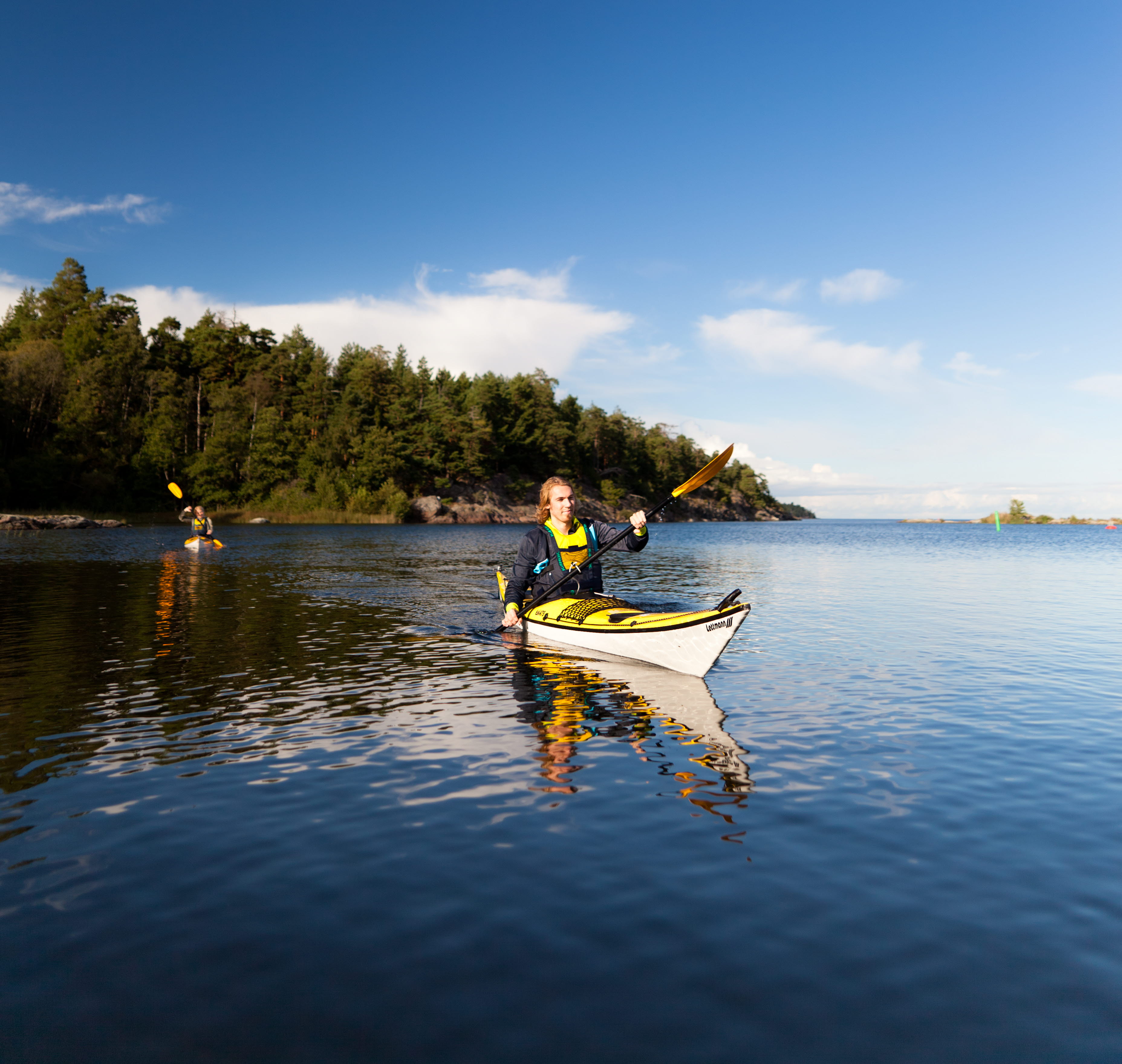 A man and a woman are paddling in their respective kayaks. The water and sky are deep blue and in the background you can see an island full of trees.