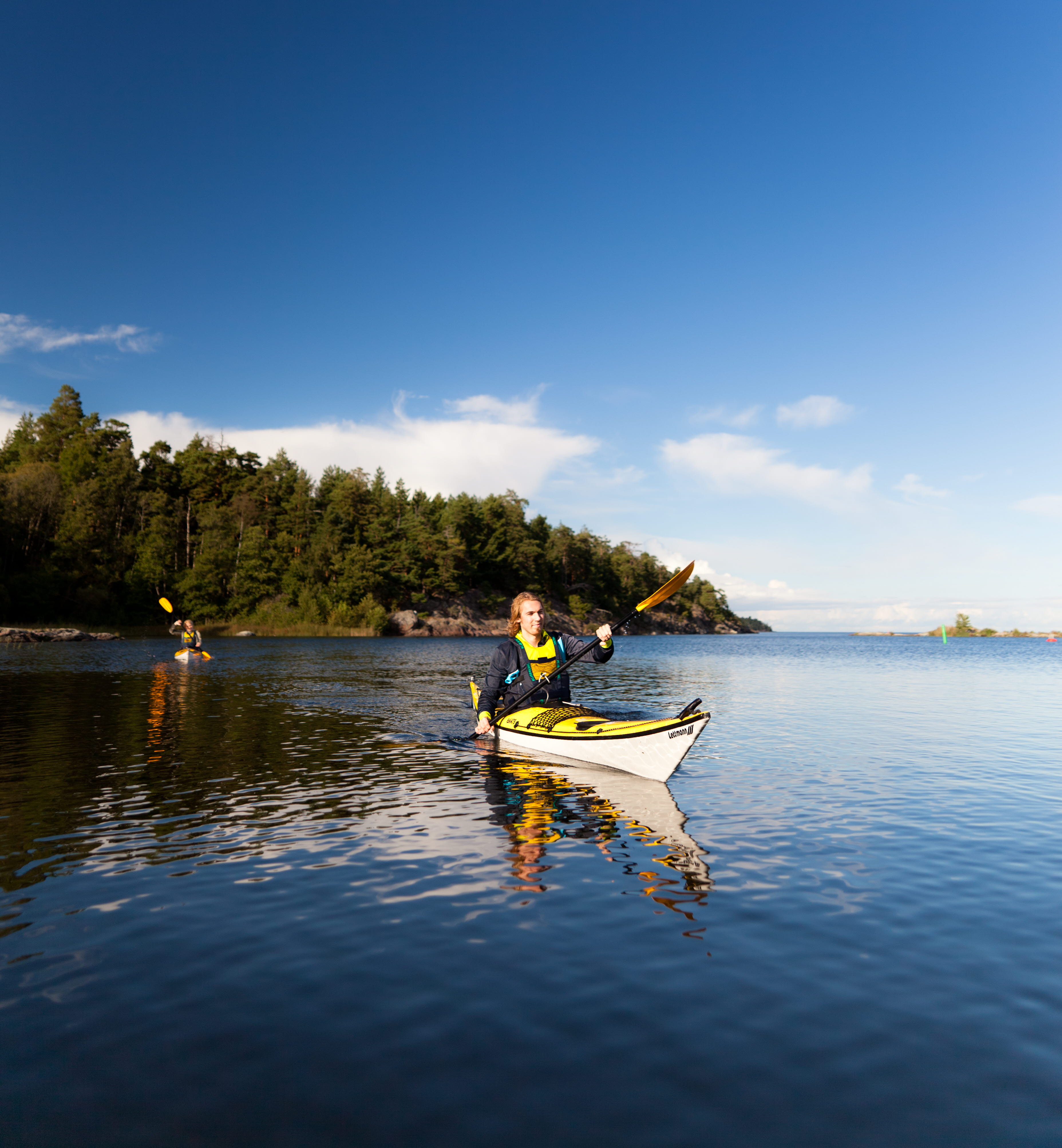 A man and a woman are paddling in their respective kayaks. The water and sky are deep blue and in the background you can see an island full of trees.