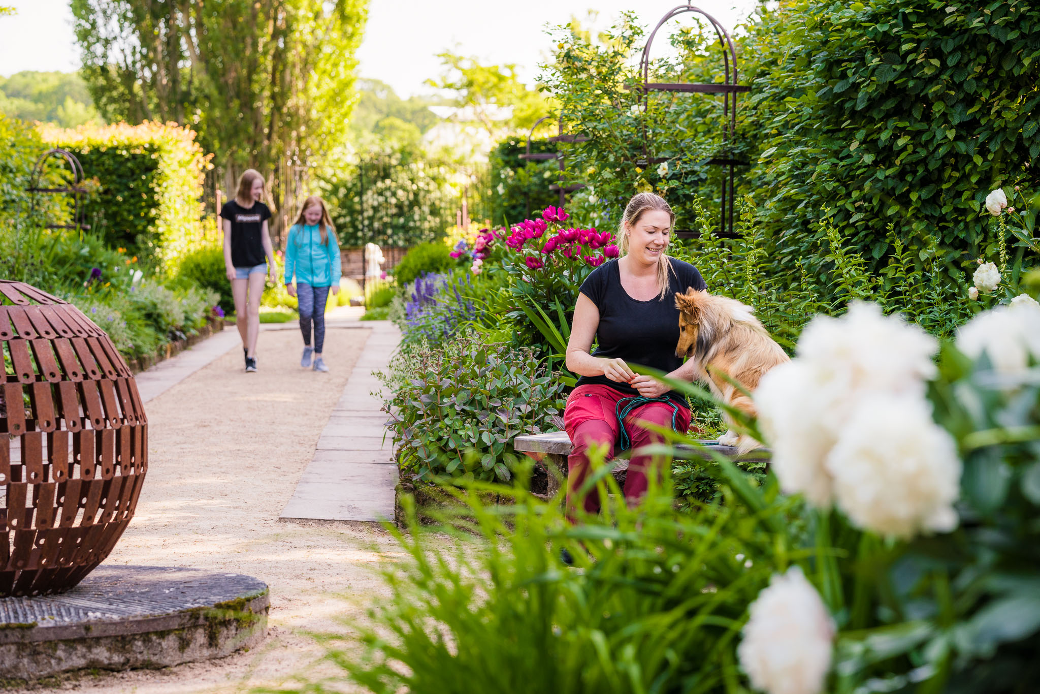 A castle garden with flowers and a walking path. Two kids are walking in the background. A woman is sitting on a bench with a dog.