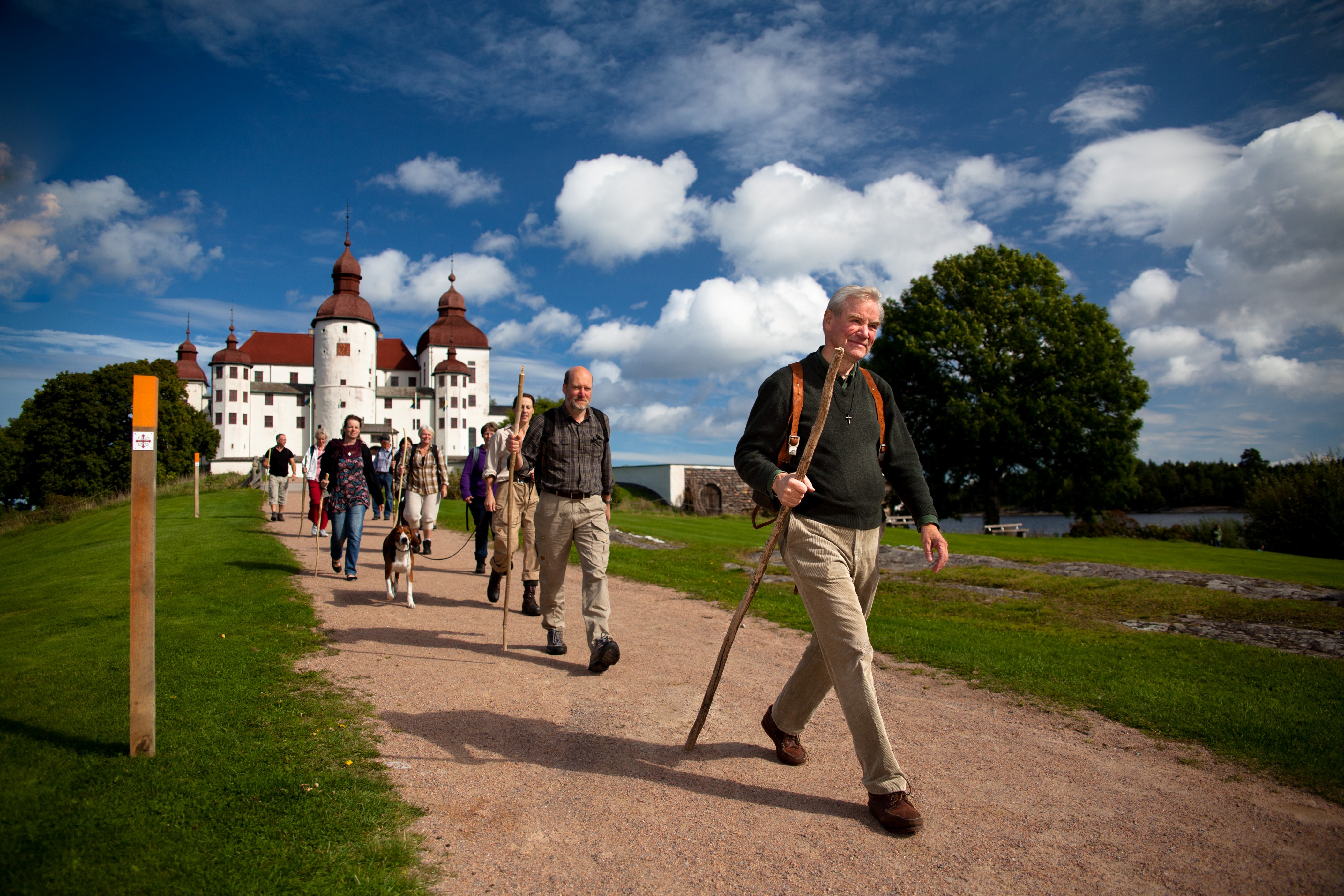 En man i förgrunden och bakom sig har hon ett led med vandrare. Stigen går utanför stora pampiga vita Läckö Slott.
