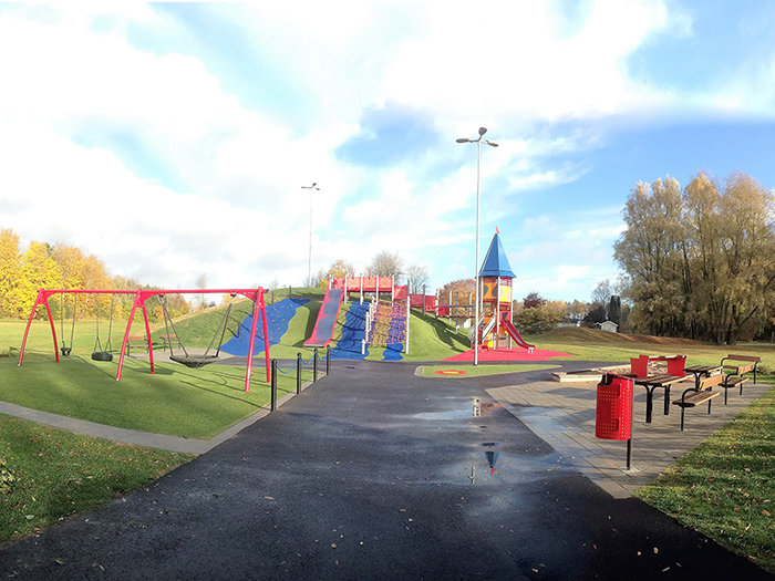 Playground placed at a hill with swings, a slide and a net to climb in. To the right are some benches and tables.