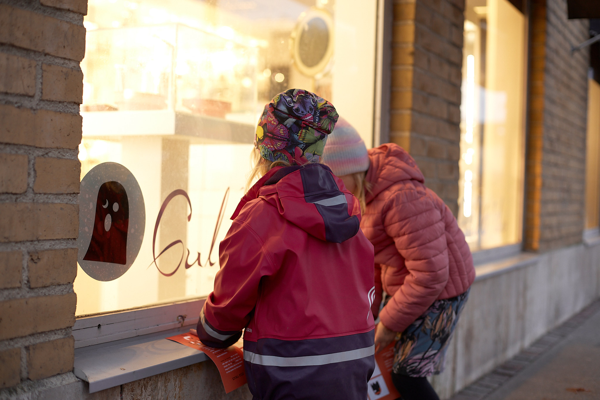 Två flickor står vid ett skyltfönster. Det sitter ett klistermärke av ett spöka i fönstret.