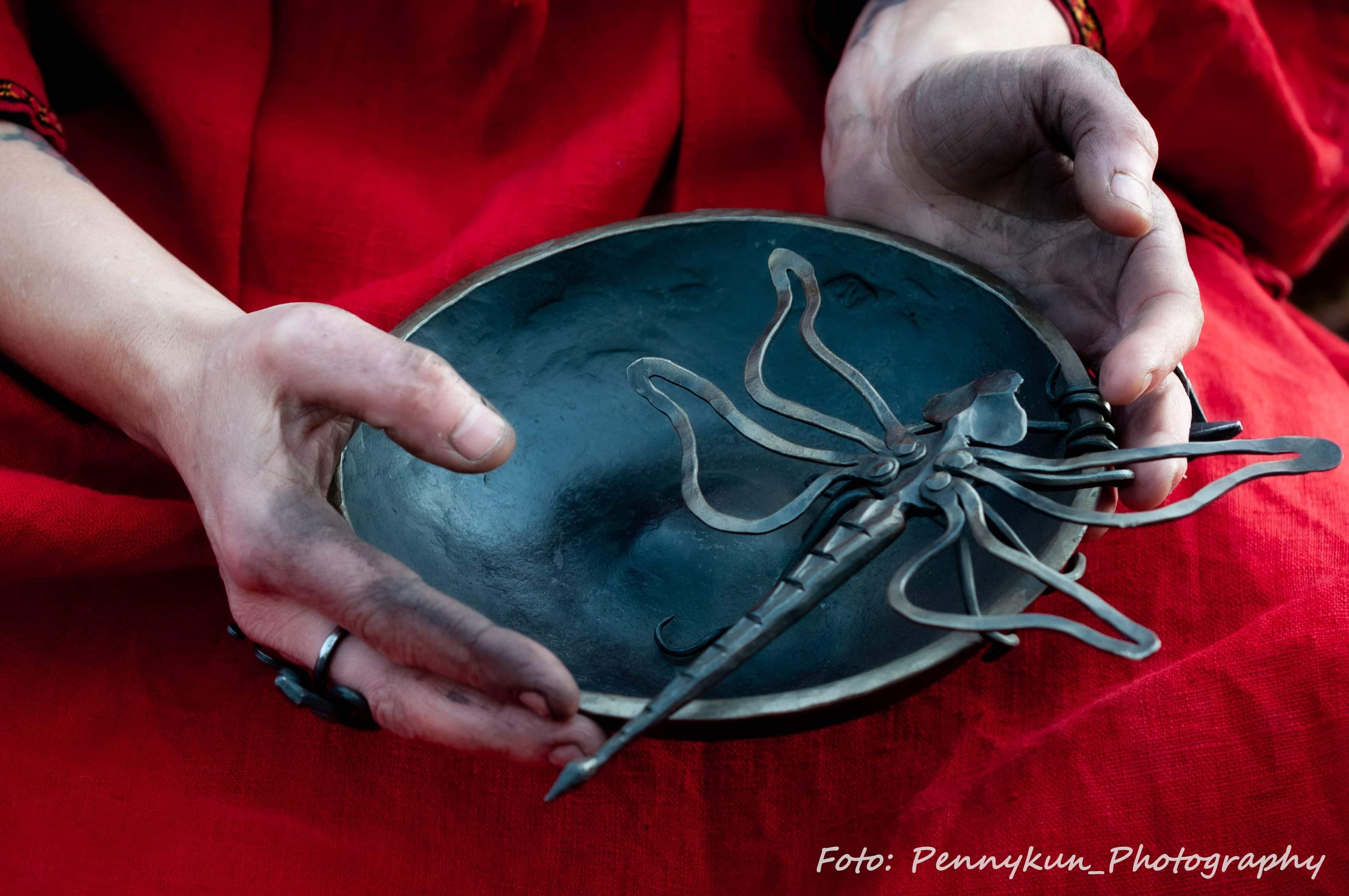 Kvinna håller i en svart skål med en slända gjort av metall i. Händerna är svarta från smide.