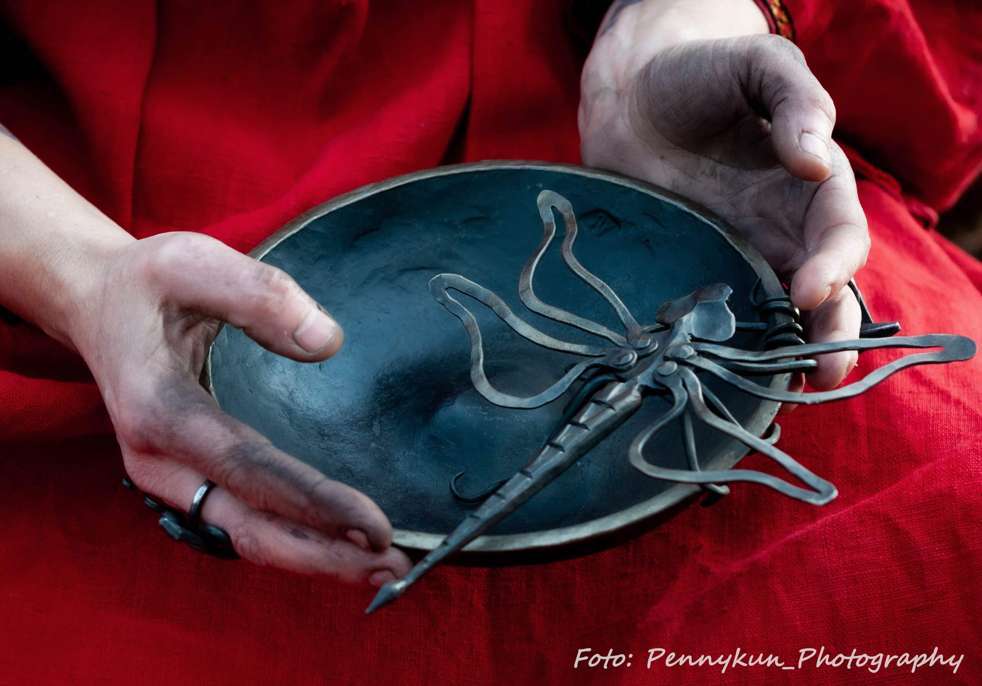 Kvinna håller i en svart skål med en slända gjort av metall i. Händerna är svarta från smide.