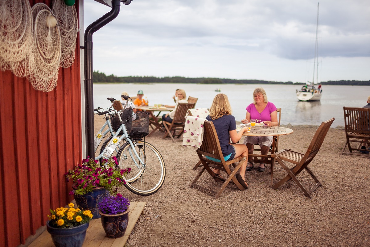 Två kvinnor sitter vid ett bord och fikar i Spikens fiskehamn. Deras cyklar står bredvid och i bakgrunden ser man Vänern och en segelbåt på väg bort från hamnen.