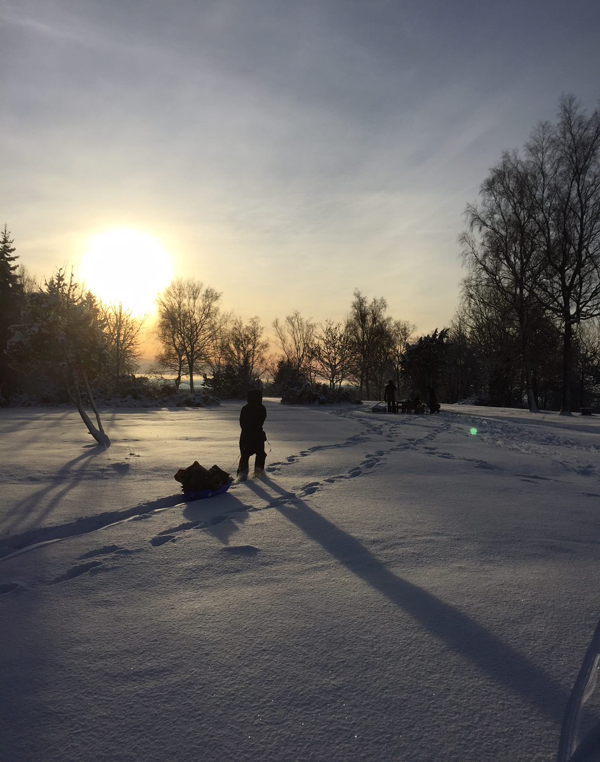 En person drar en pulka efter sig i ett snölandskap. Solen står lågt på himlen. 