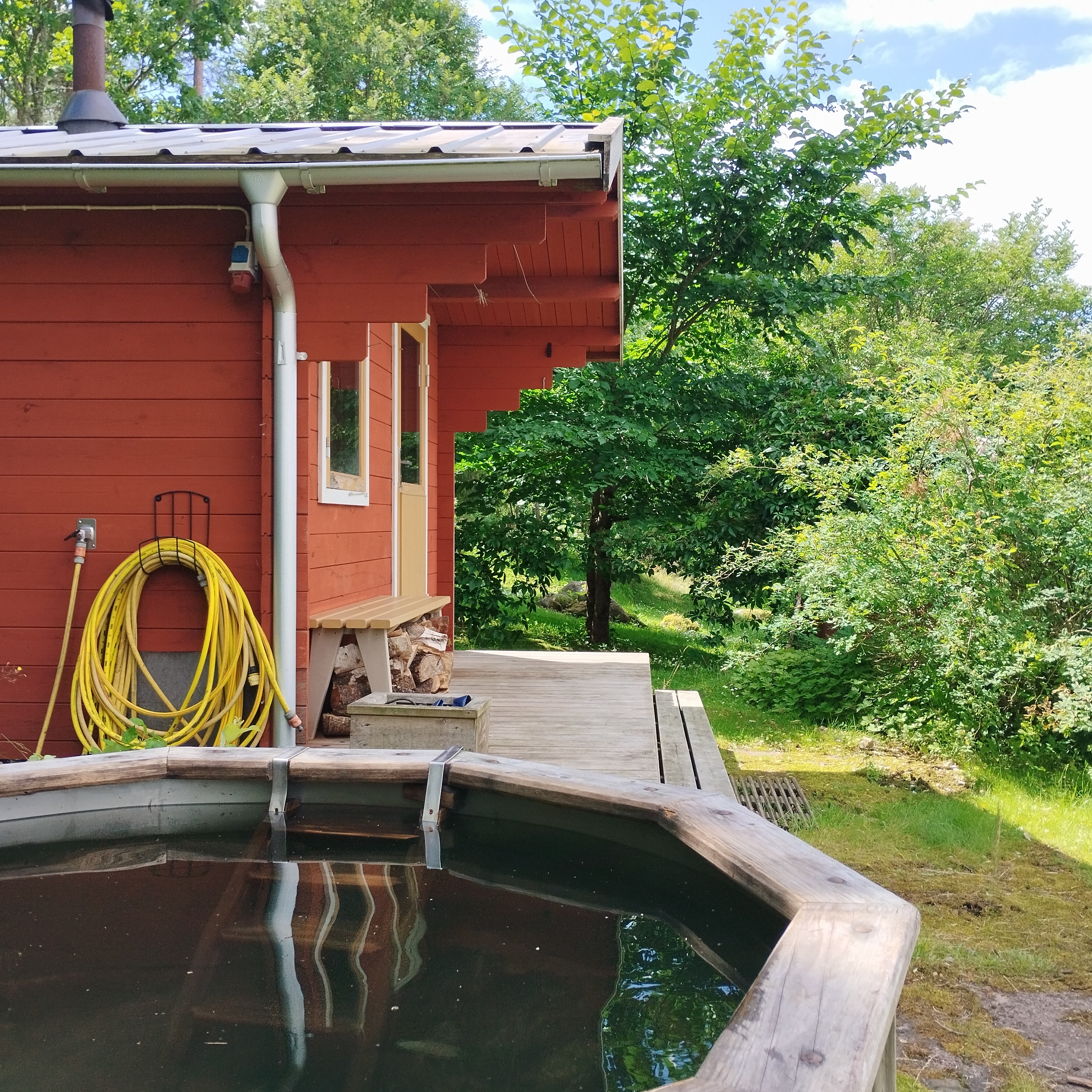 Röd liten stuga omgiven av grönska. Bredvid huset finns en kall badrtunna,.