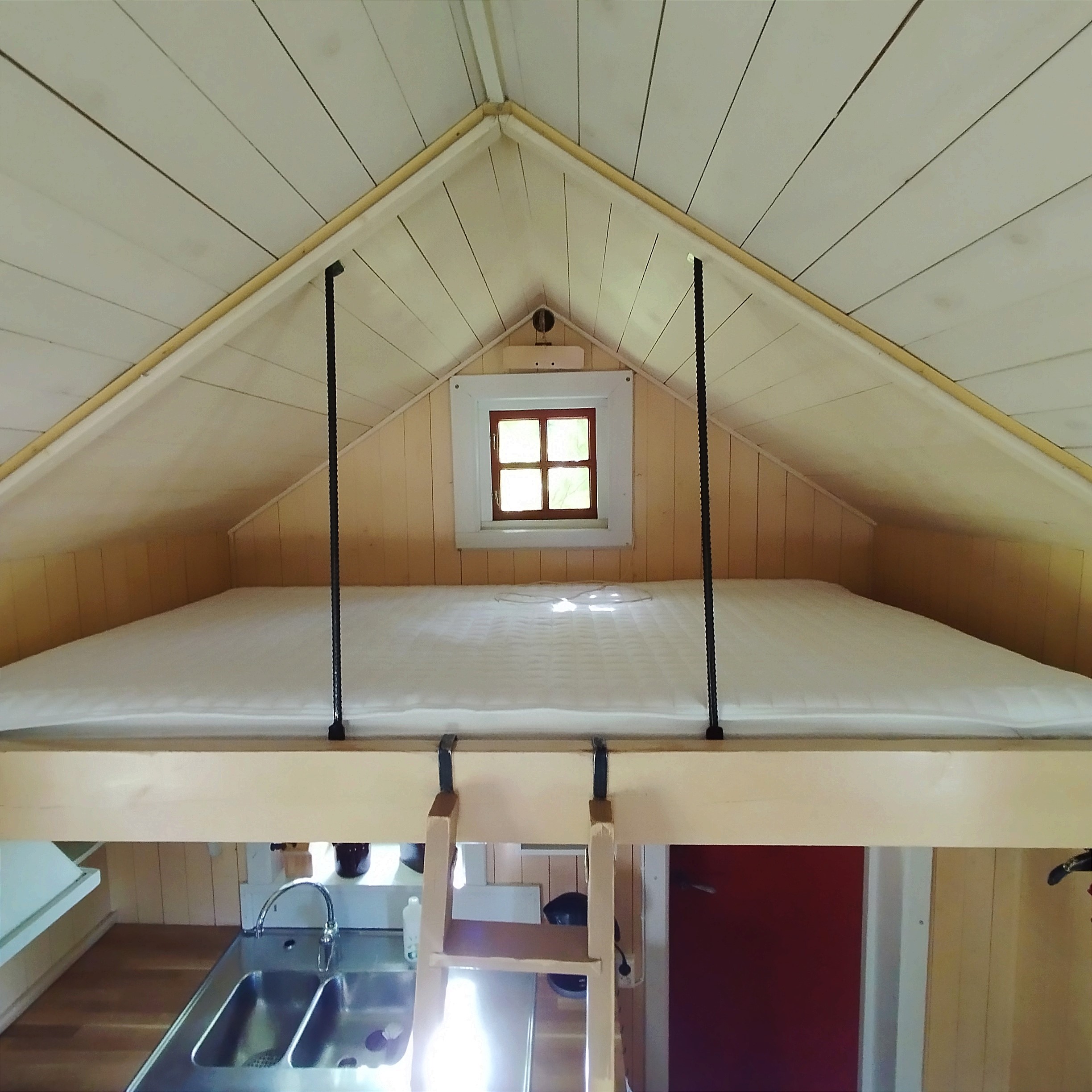 Liten stuga med loft med stor bäddmadrass. Nedanför loftet skymtas ett pentry.