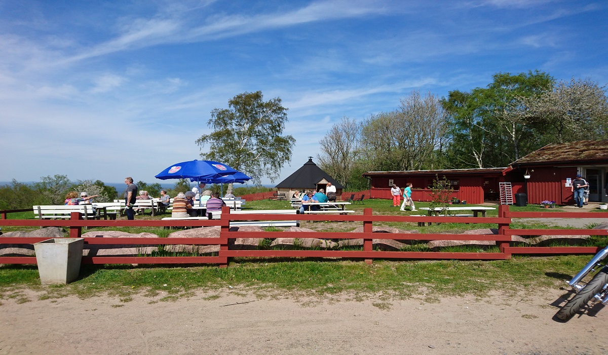 En plats utomhus med träd i bakgrunden. Ett rött staket avskärmar en fikaplats med bord och stolar.