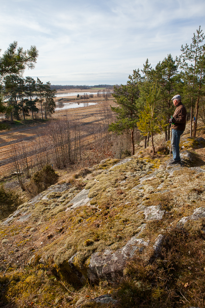 En äldre man står på en mossklädd bergkulle och ser ut över åker och vatten.