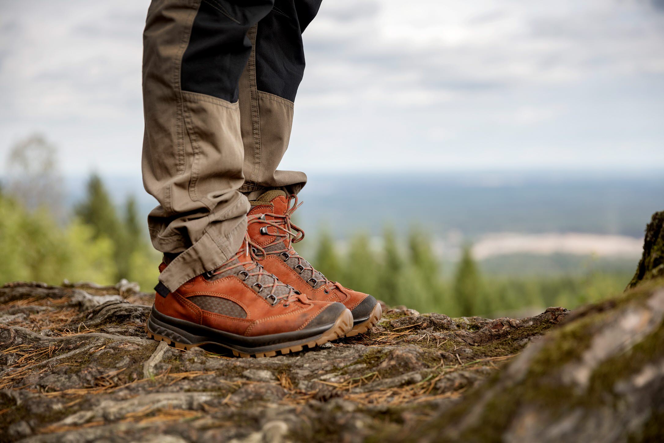 I bild syns en persons byxor och vandringskängor. Personen står på en höjd med en utsikt i bakgrunden.