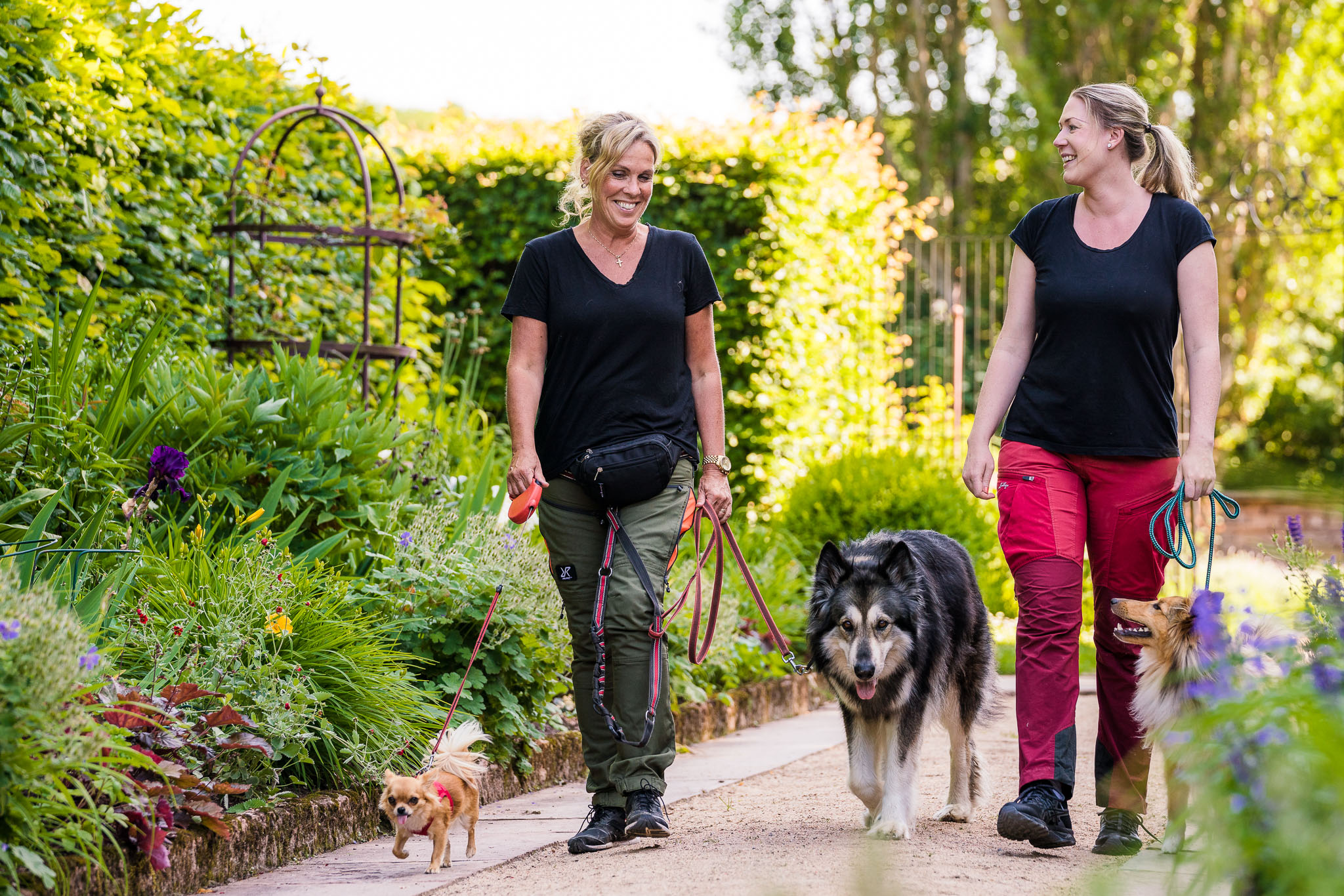 Två kvinnor med hundar iklädda vandringskläder i en vacker trädgårdsmiljö.