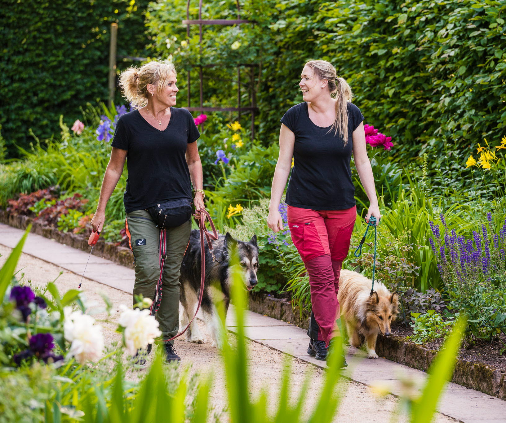 Två kvinnor med hundar iklädda vandringskläder i en vacker trädgårdsmiljö.