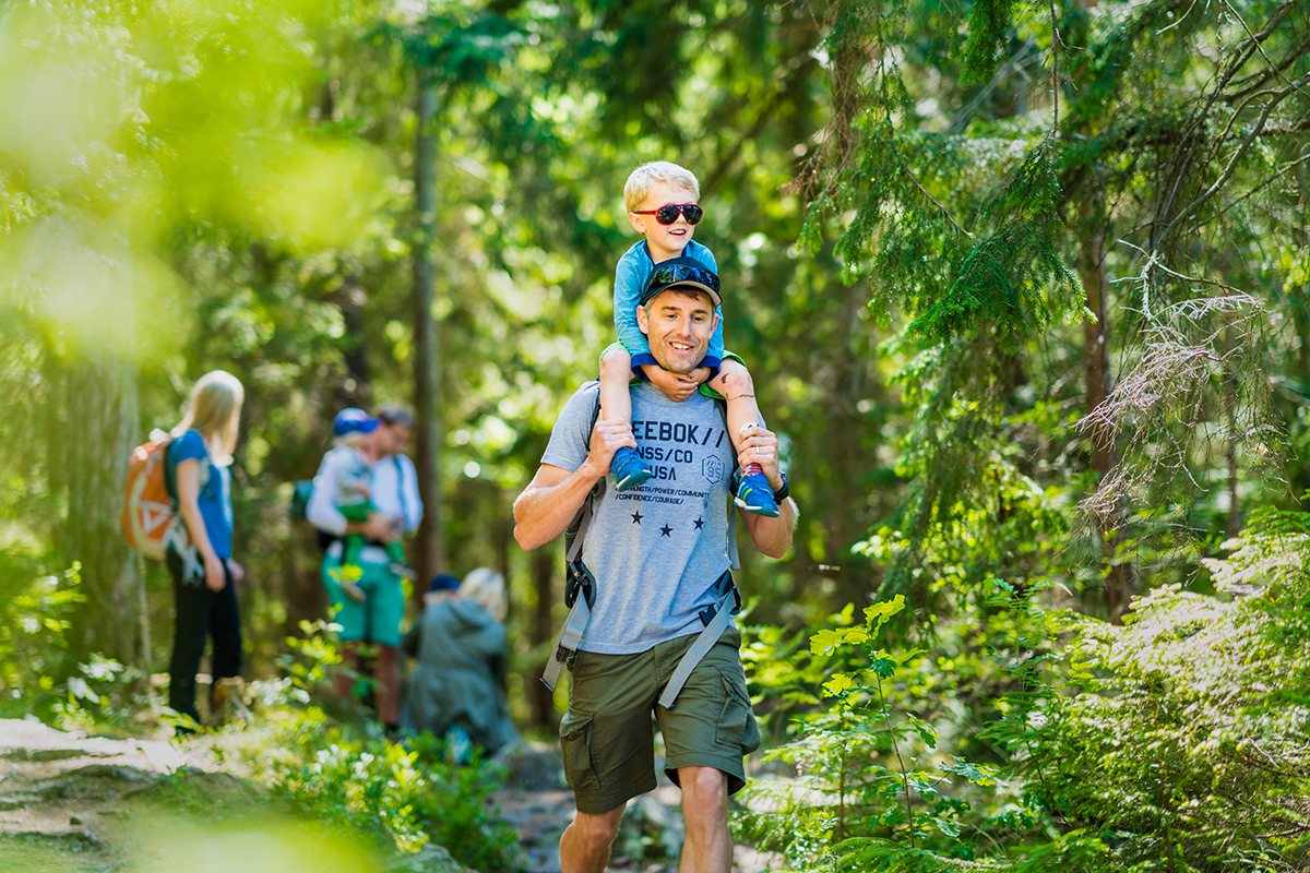 A father with his son on his shoulders in lush green forest. In the background, the rest of the hiking community is spotted.