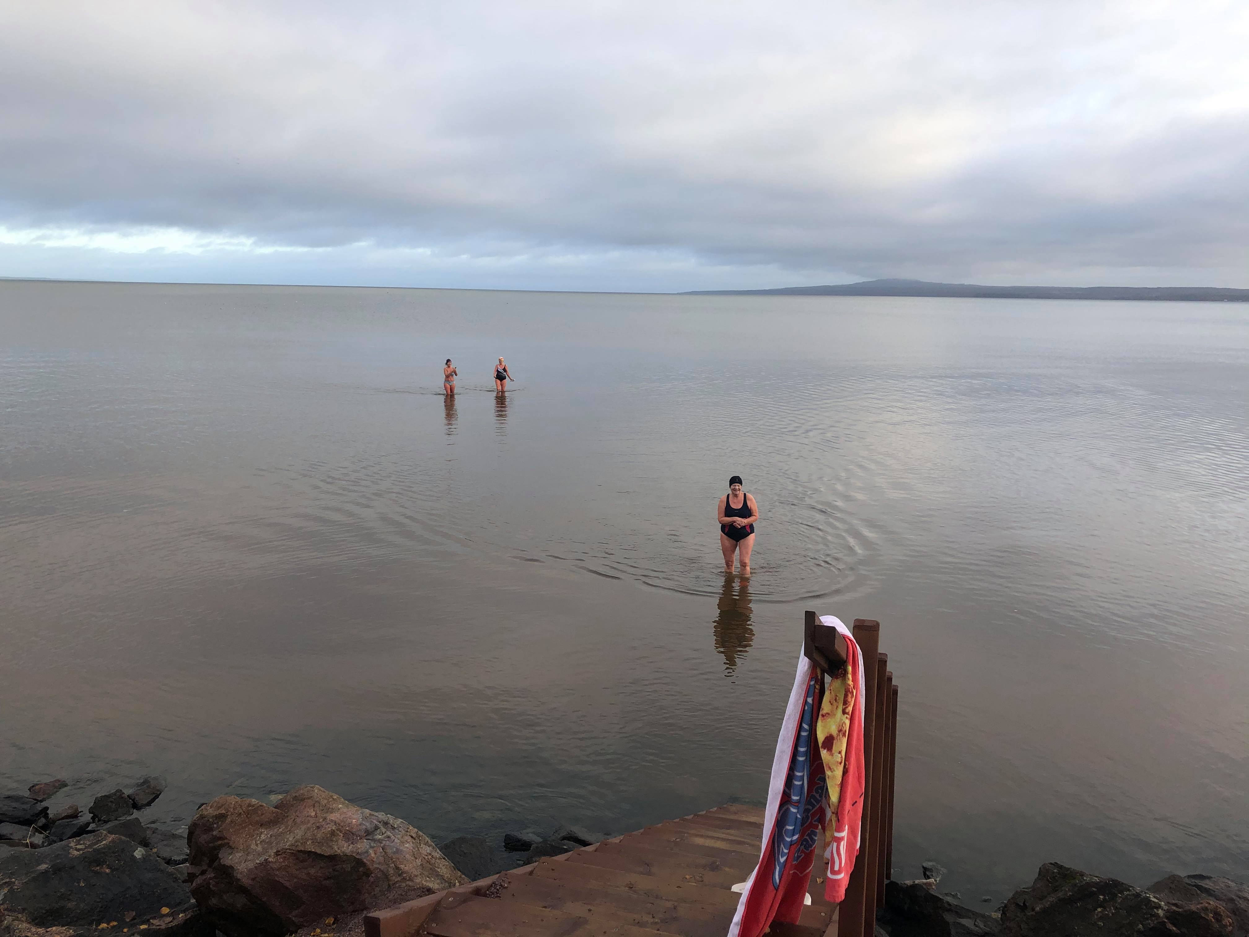 Tre kvinnor som vinterbadar. På bryggan hänger en handduk. I bakgrunden skymtar Kinnekulle.