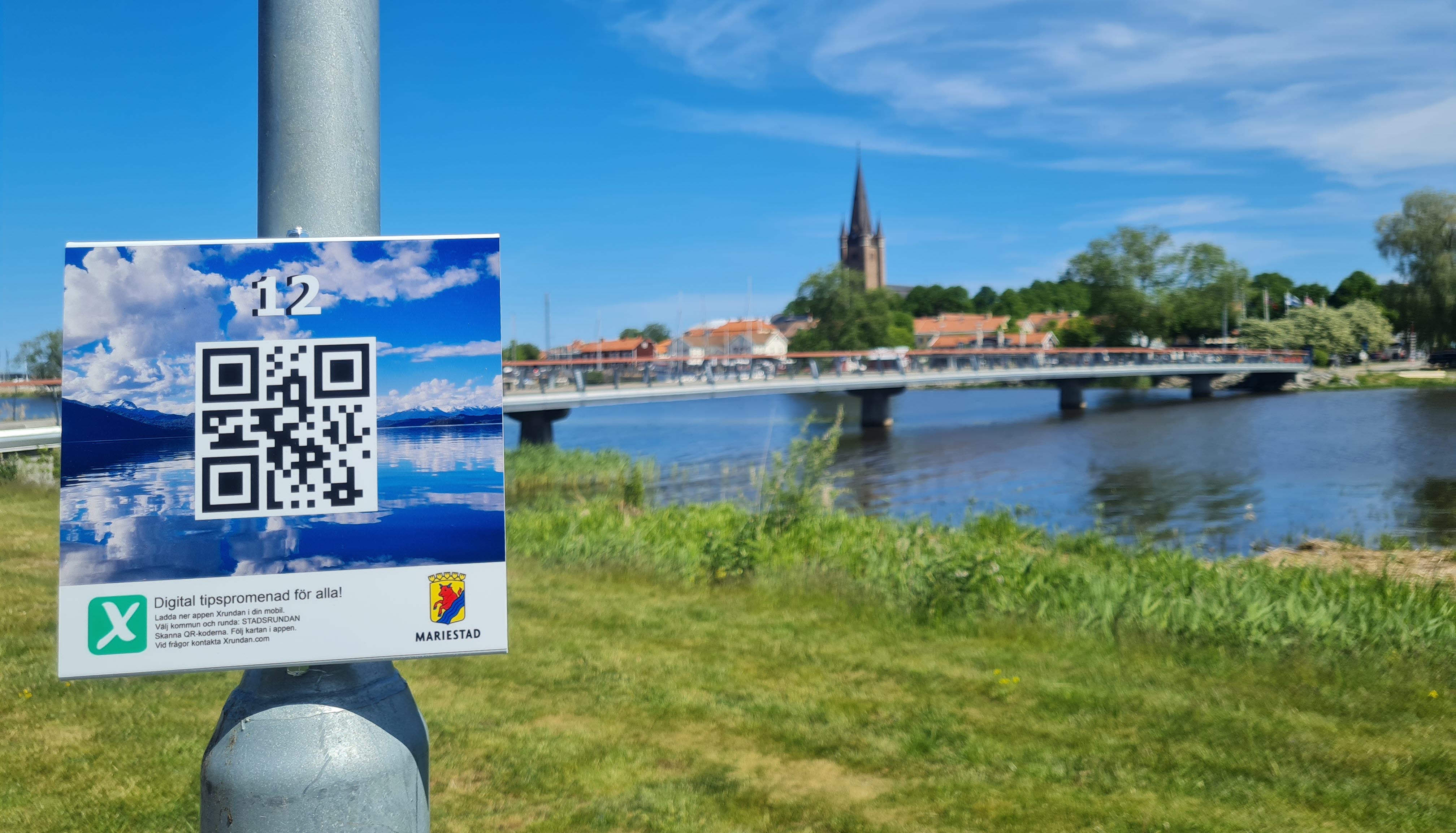 Xrundan skylt på en lyktstolpe i förgrunden. Gräs, vatten, en bro, hustak och ett kyrktorn i bakgrunden.