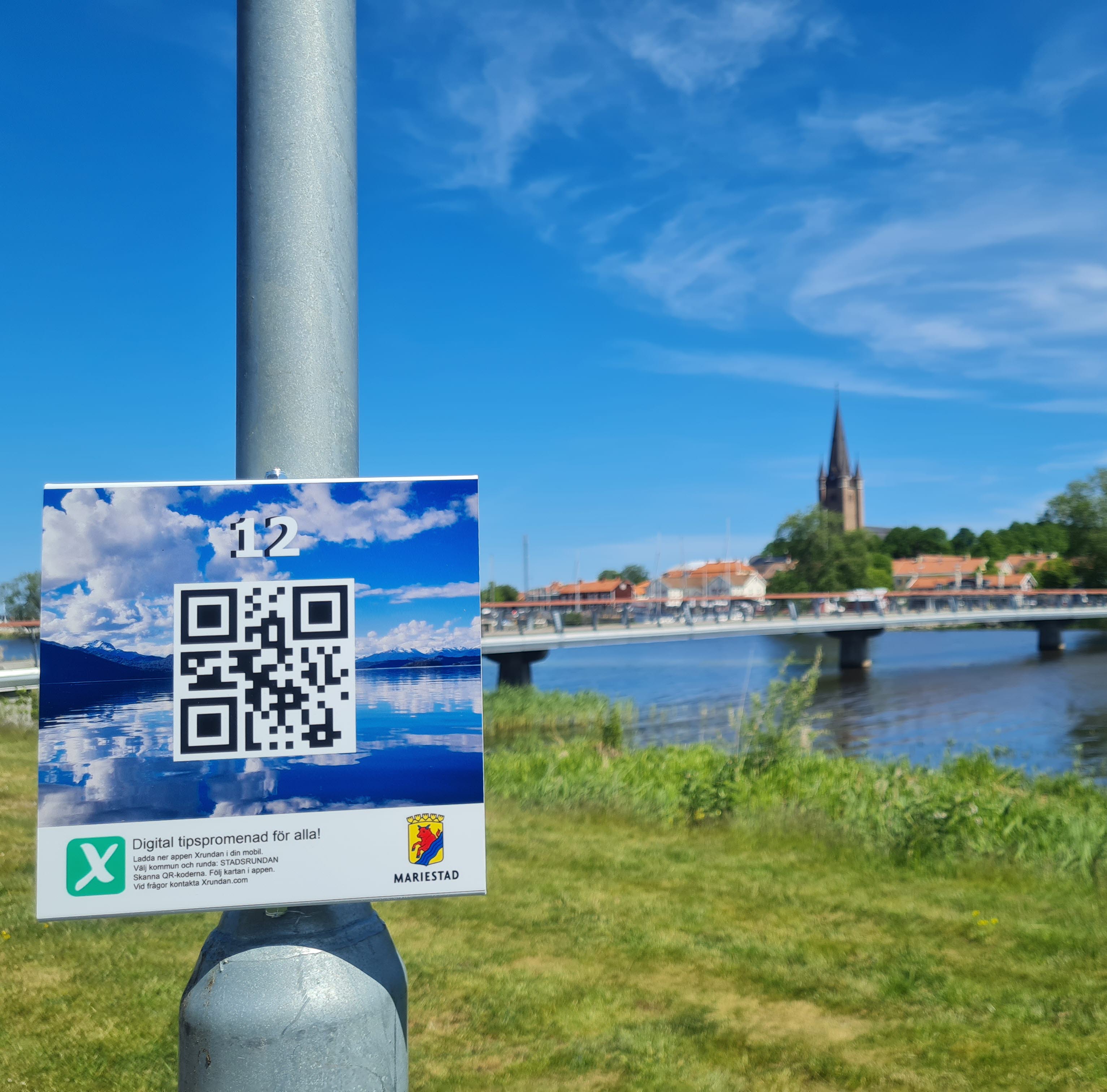 Xrundan skylt på en lyktstolpe i förgrunden. Gräs, vatten, en bro, hustak och ett kyrktorn i bakgrunden.