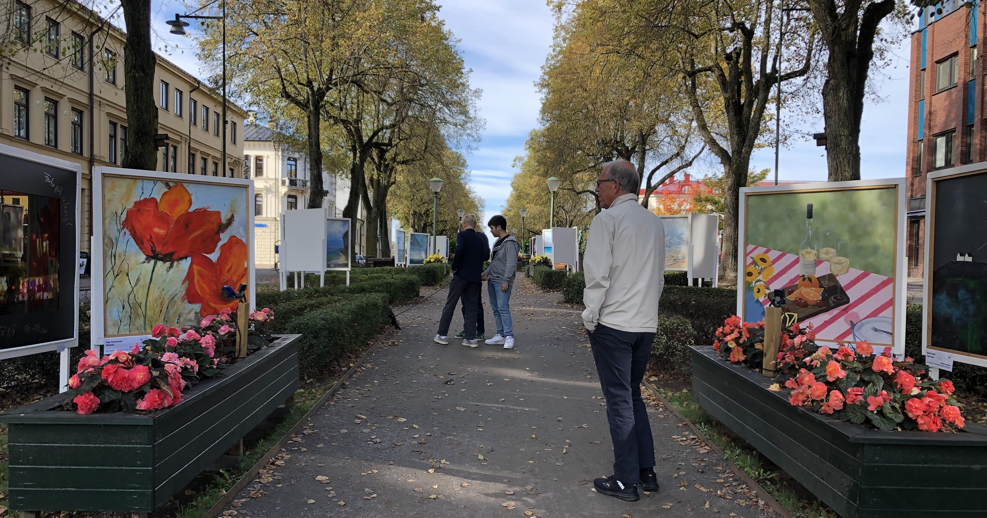Tavlor utomhus i en trädallé i stadsmijö. 4 personer tittar på tavlorna.