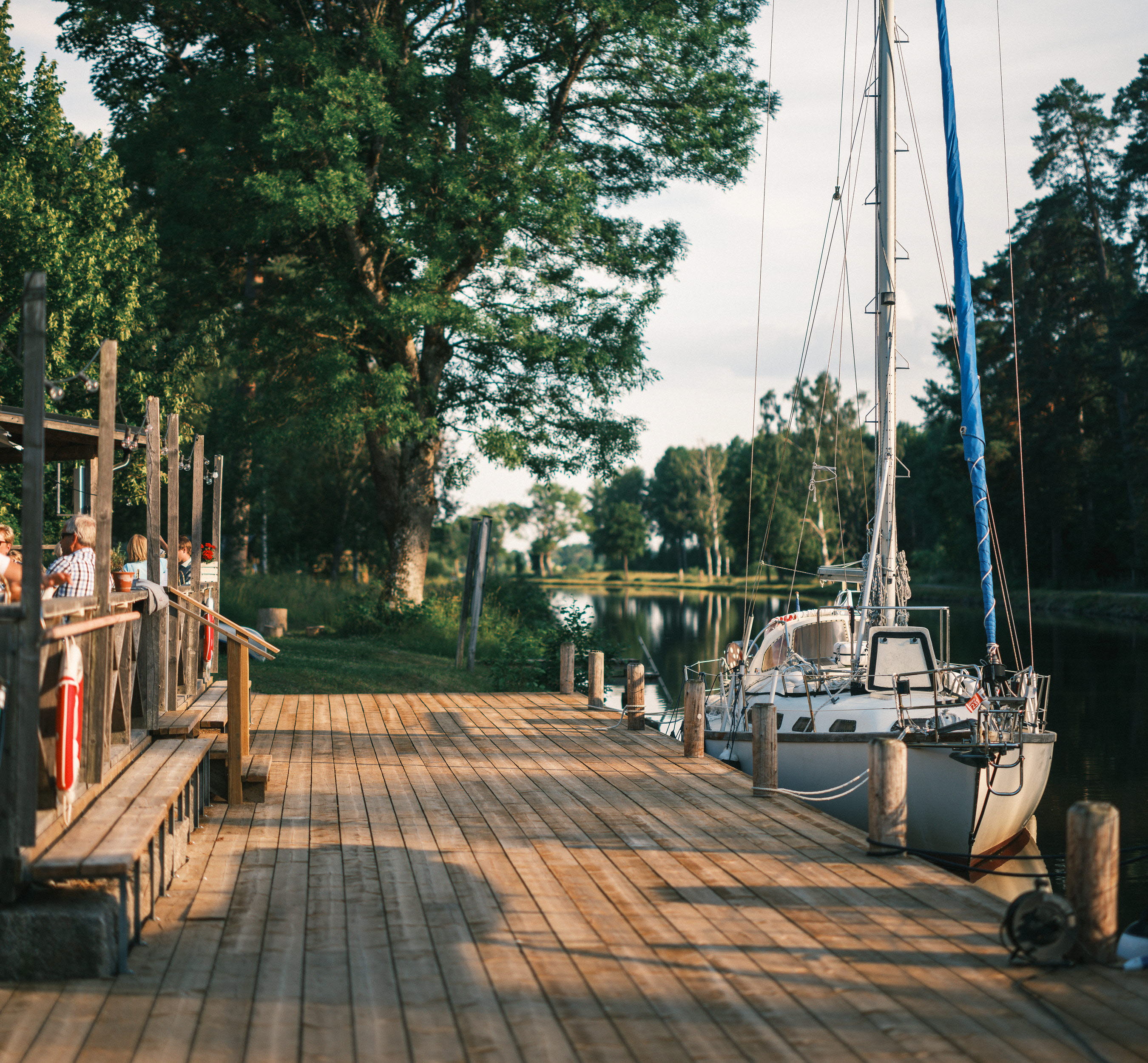 En brygga vid Göta kanal, en segelbåt och människor på en uteservering.