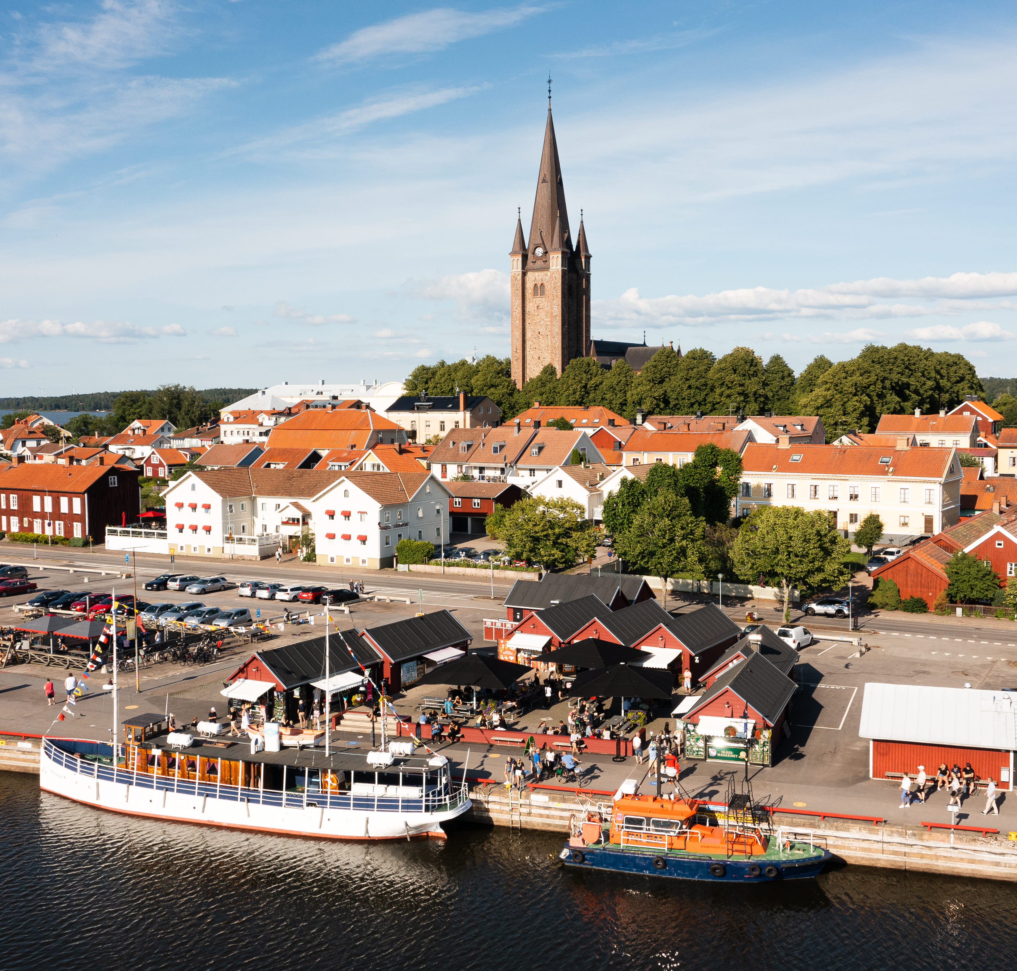 Flygfoto över bodarna i Mariestads gästhamn. Del av Gamla Stan med domkyrkan. Båtar i vattnet i förgrunden.