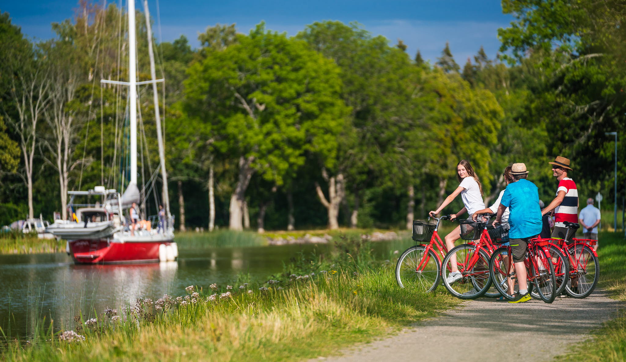 Tre personer stå med sina cyklar på kanalbanken och tittar på en segelbåt i Göta kanal.