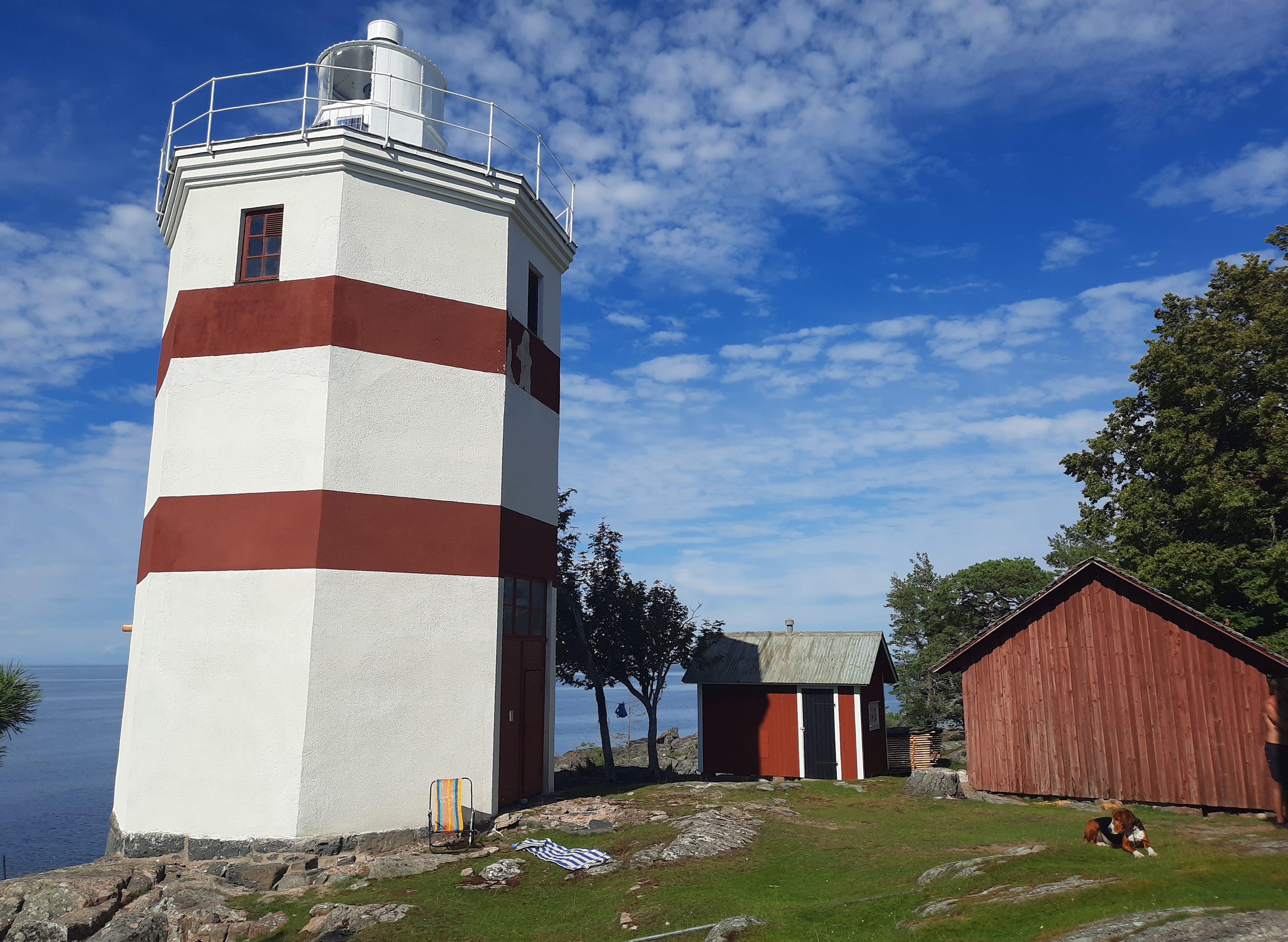 Åttakantig fyr i vitt med två röda horisontala streck på en klippa med Vänern bakom och två röda låga uthus intill.