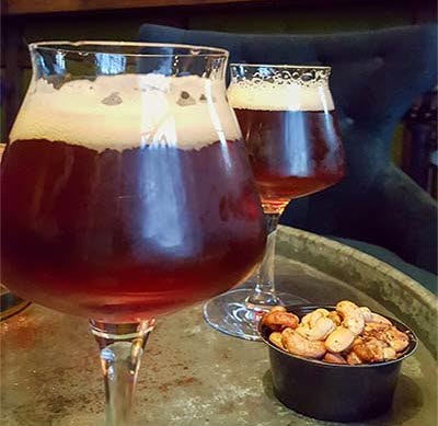 Två glas öl och en skål med nötter på ett bord med bardisk i bakgrunden.