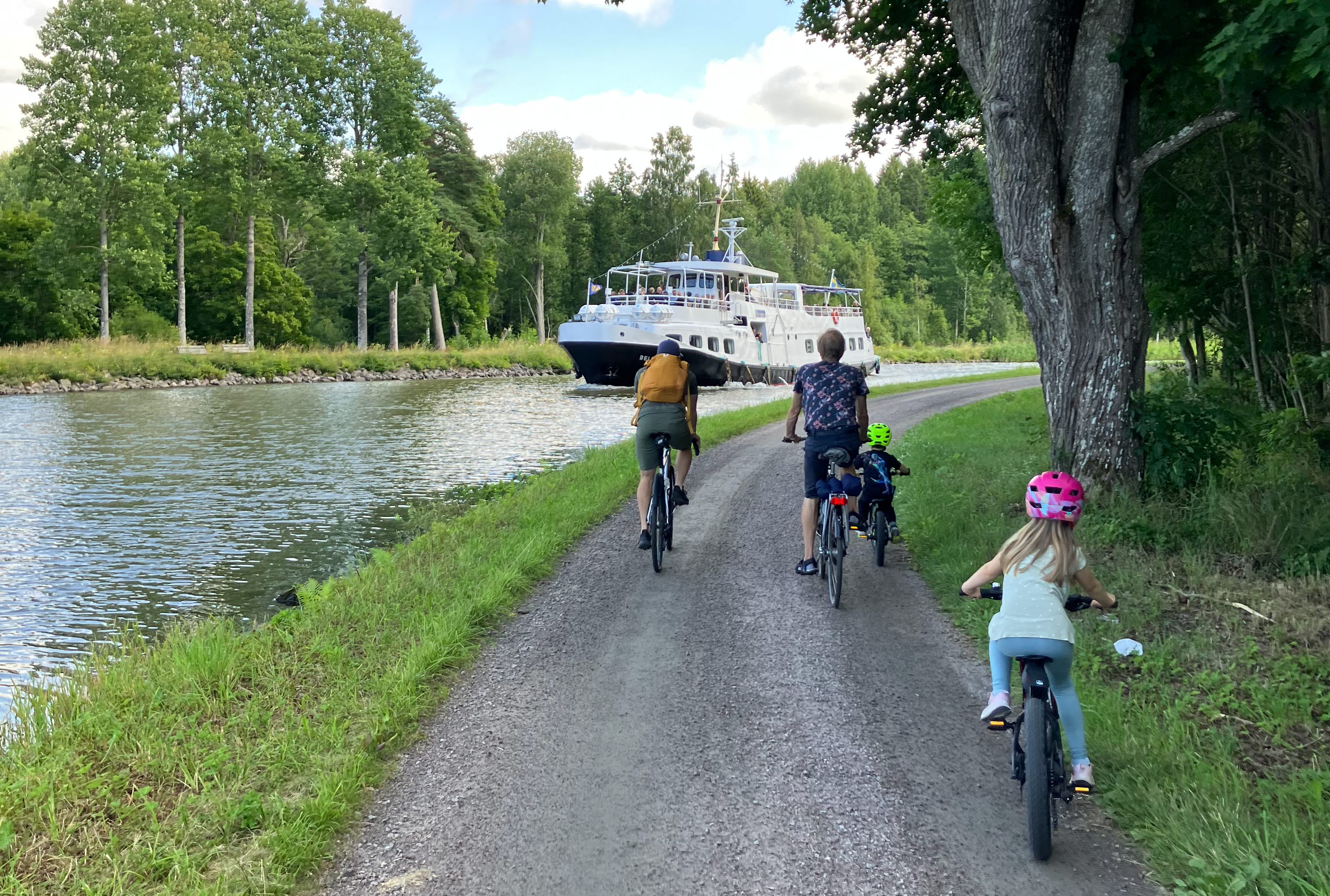 2 vuxna och 2 barn cyklar på en liten grusväg intill Göta kanal. På kanalen syns passagerarbåten M/S Bellevue.