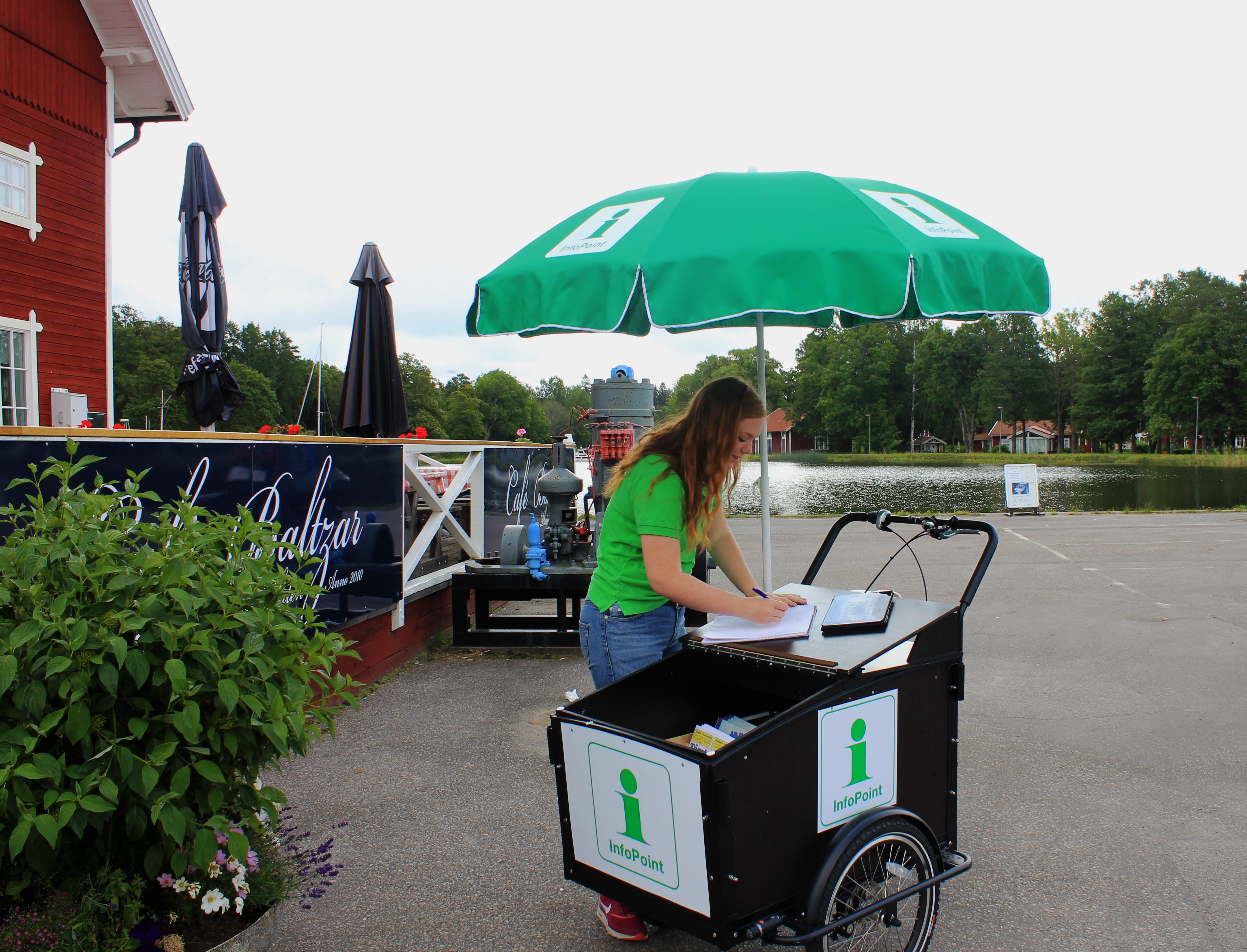 En packcykel med broschyrer, ett grönt parasoll och en kvinna i grön tröja står utanför en uteservering vid Göta kanal.