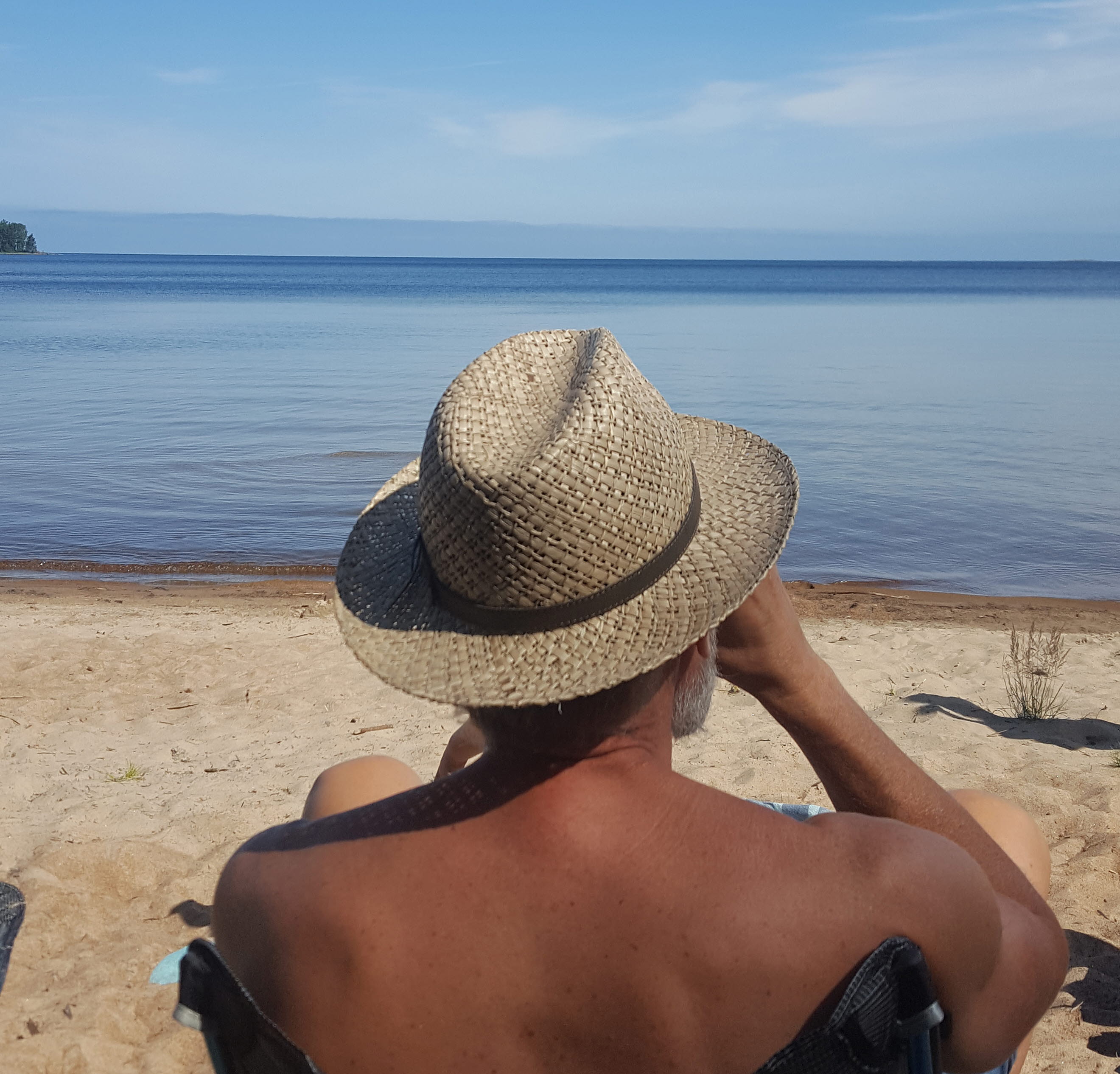 Man i hatt sitter i solstol och dricker kaffe på badstranden Store Vite Sand på Brommö i Vänern. 