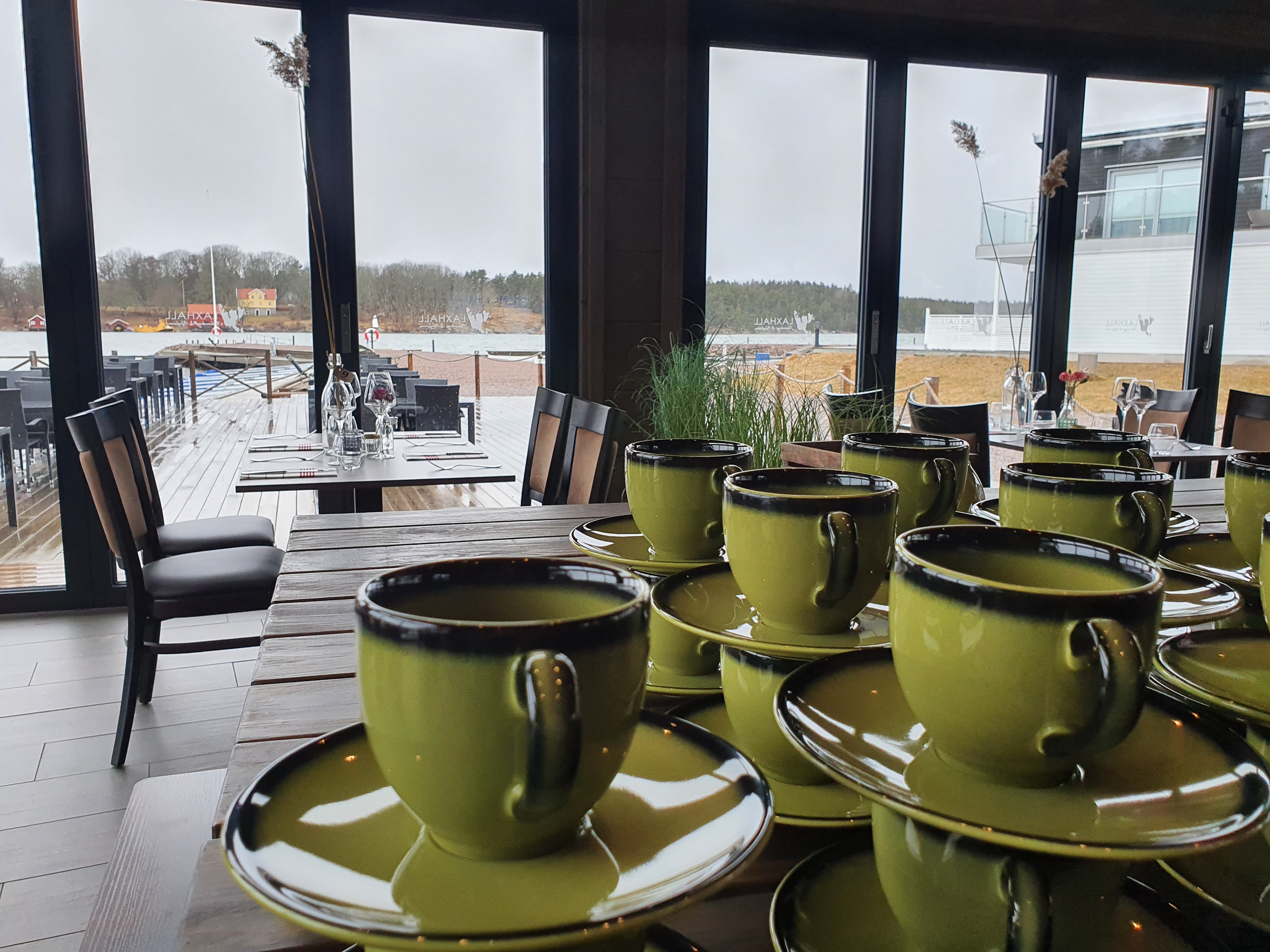 Gröna kaffekoppar i förgrunden och dukade bord med utsikt över sjö.