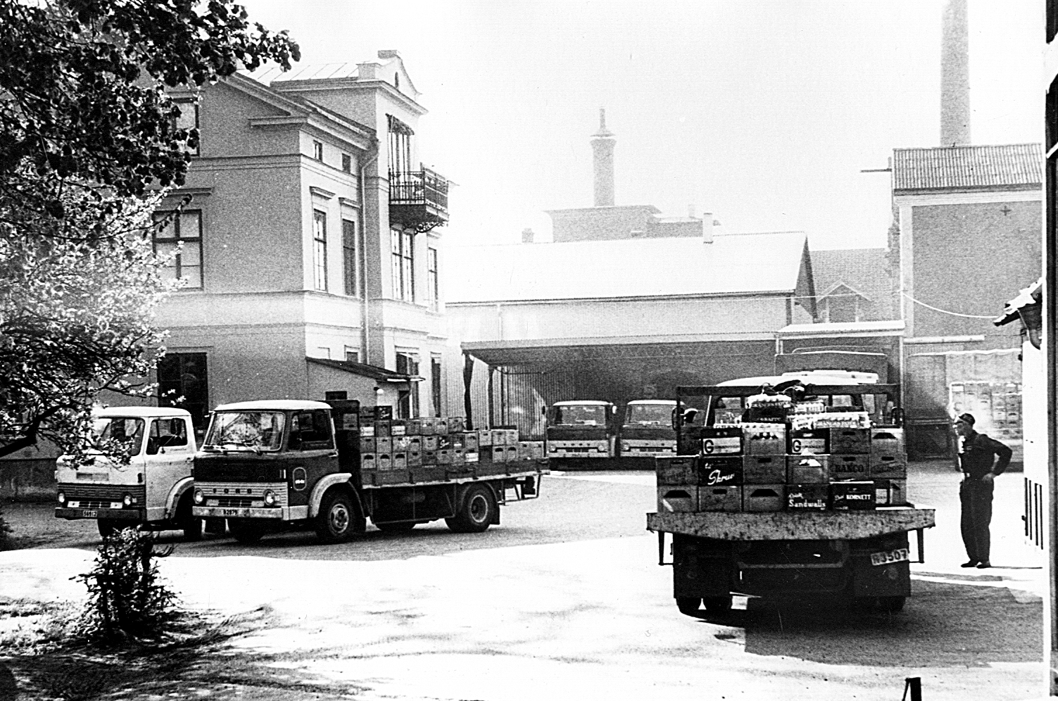 Svartvit historisk bild på Mariestads bryggeri, ett torn med skorstenar och lastbilar förgrunden. 