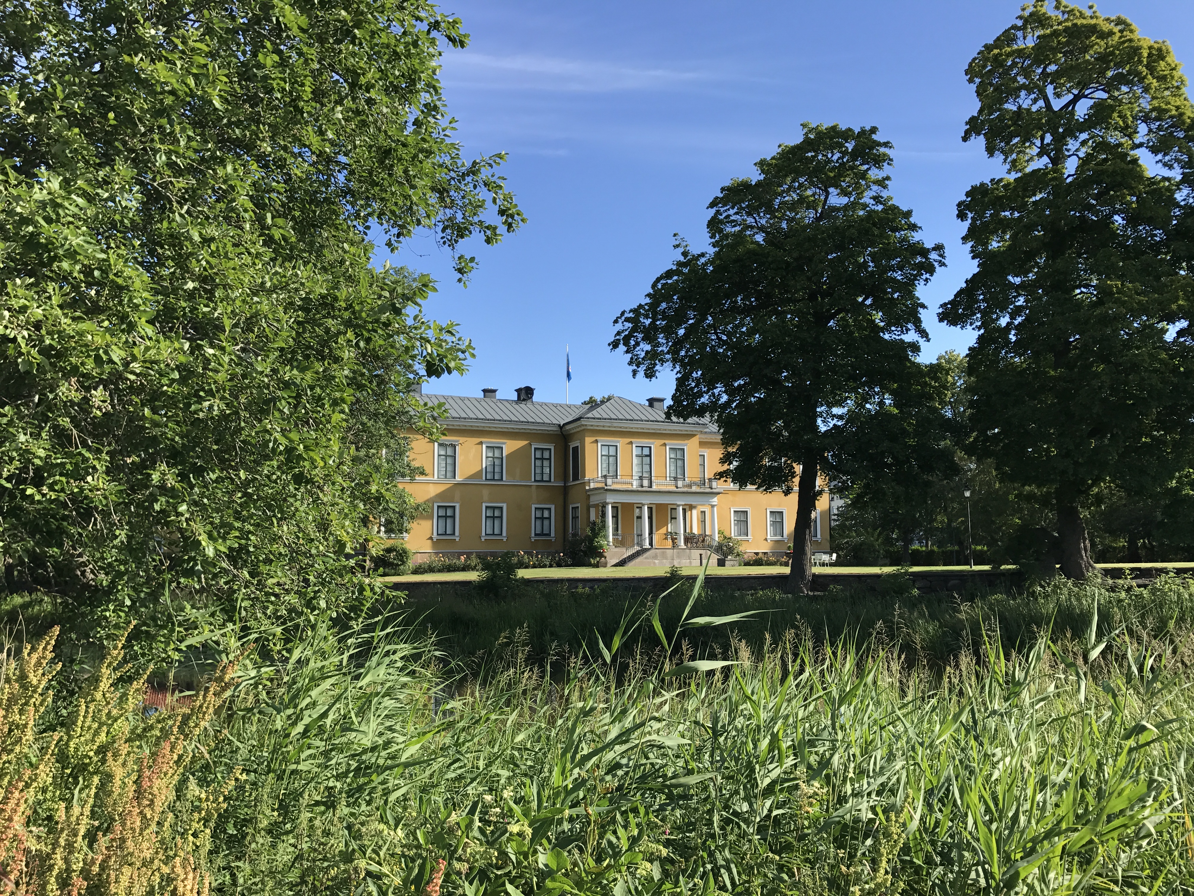 Ett stort gult hus med många fönster, träd och grön vass i förgrunden.