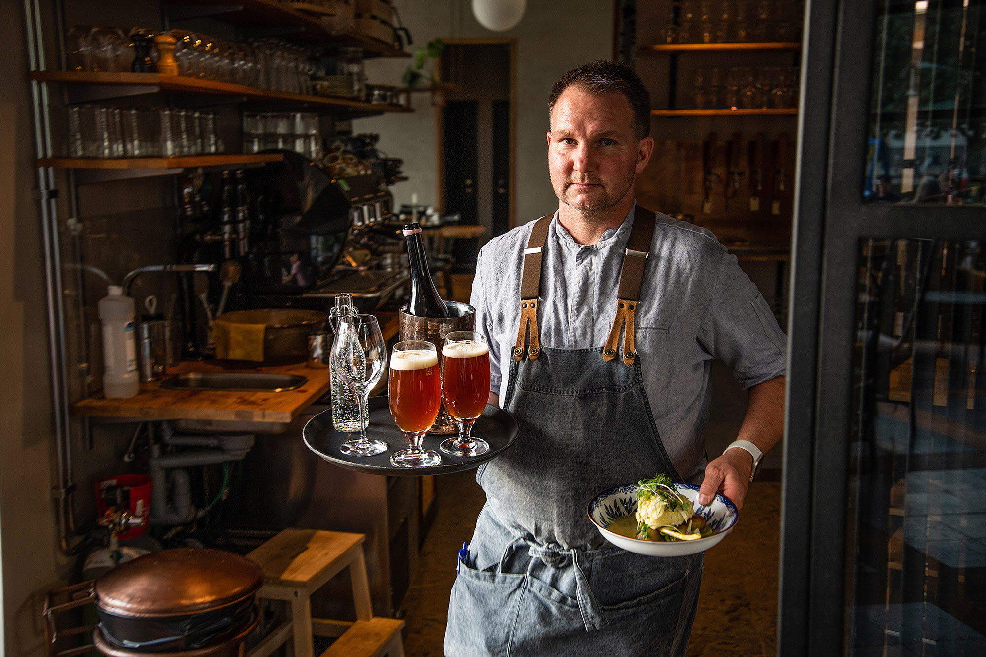 Waiter on his way out of the restaurant with a plate of food and a tray of filled beer glasses.