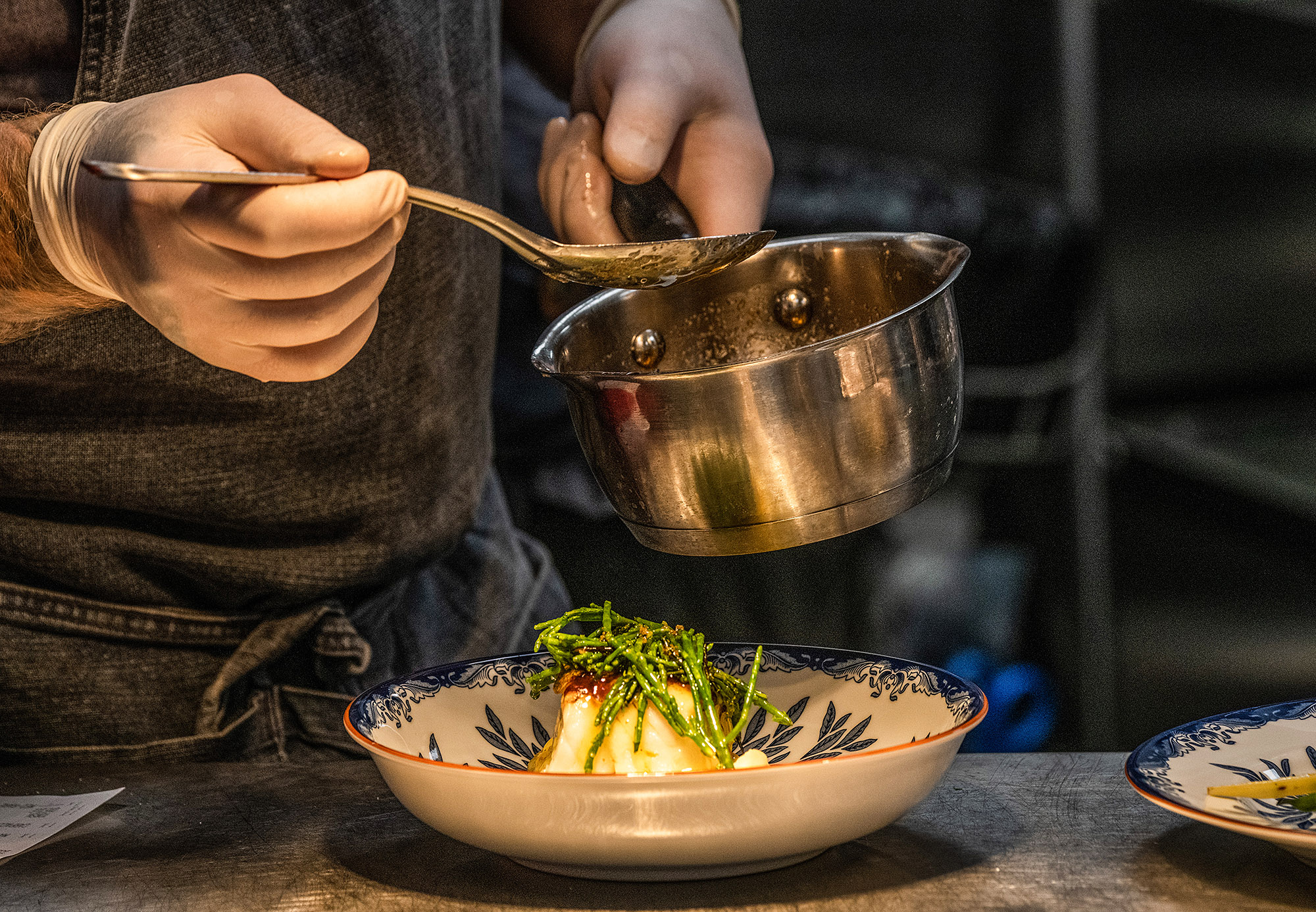 Uppläggning av maträtt på tallrik, kastrull och såssked hålls av ett par händer. 