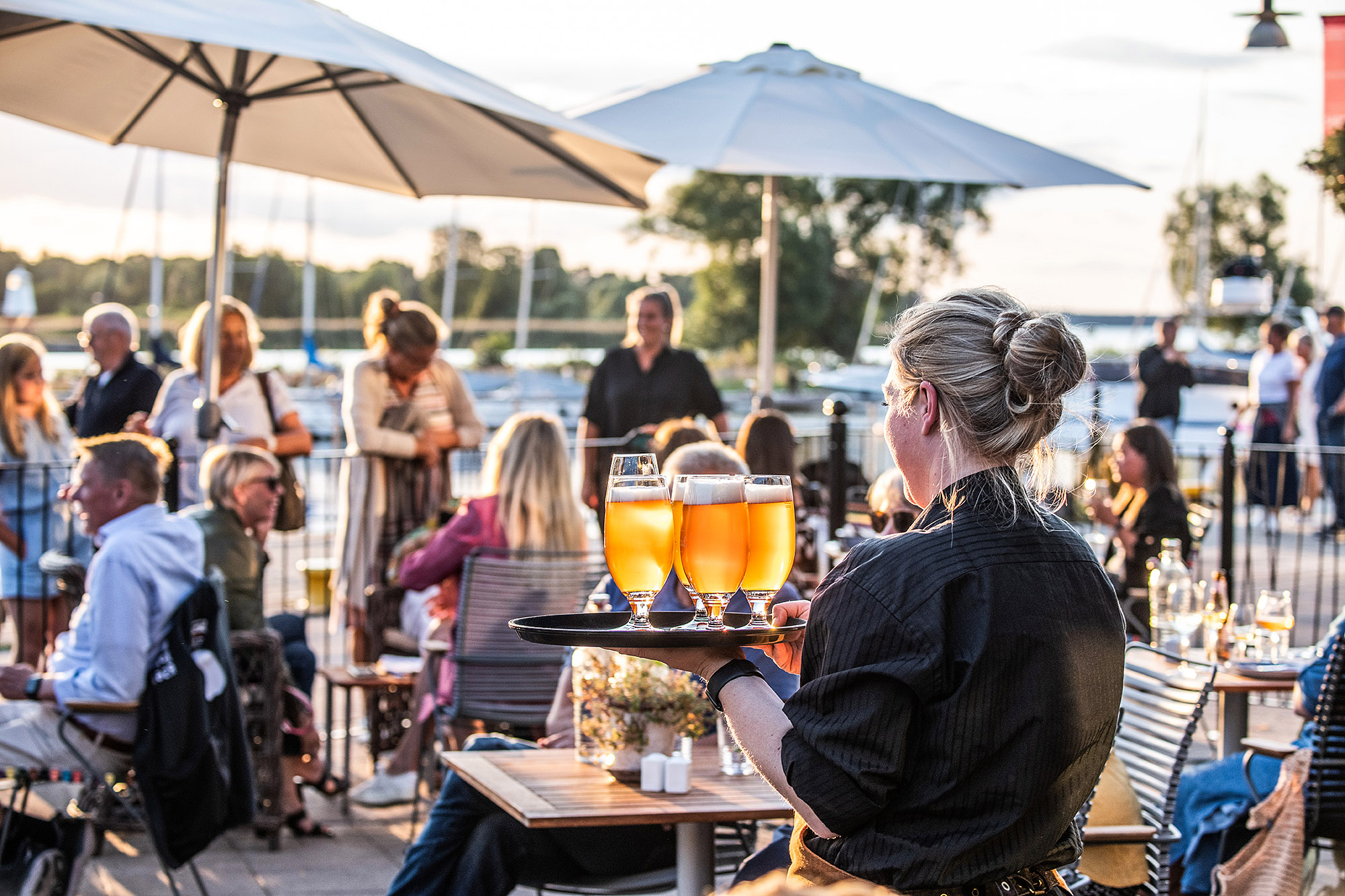Ryggtavlan på en servitris bärande på en bricka fyllda ölglas på väg mot gästerna på uteservering med parasoller. 
