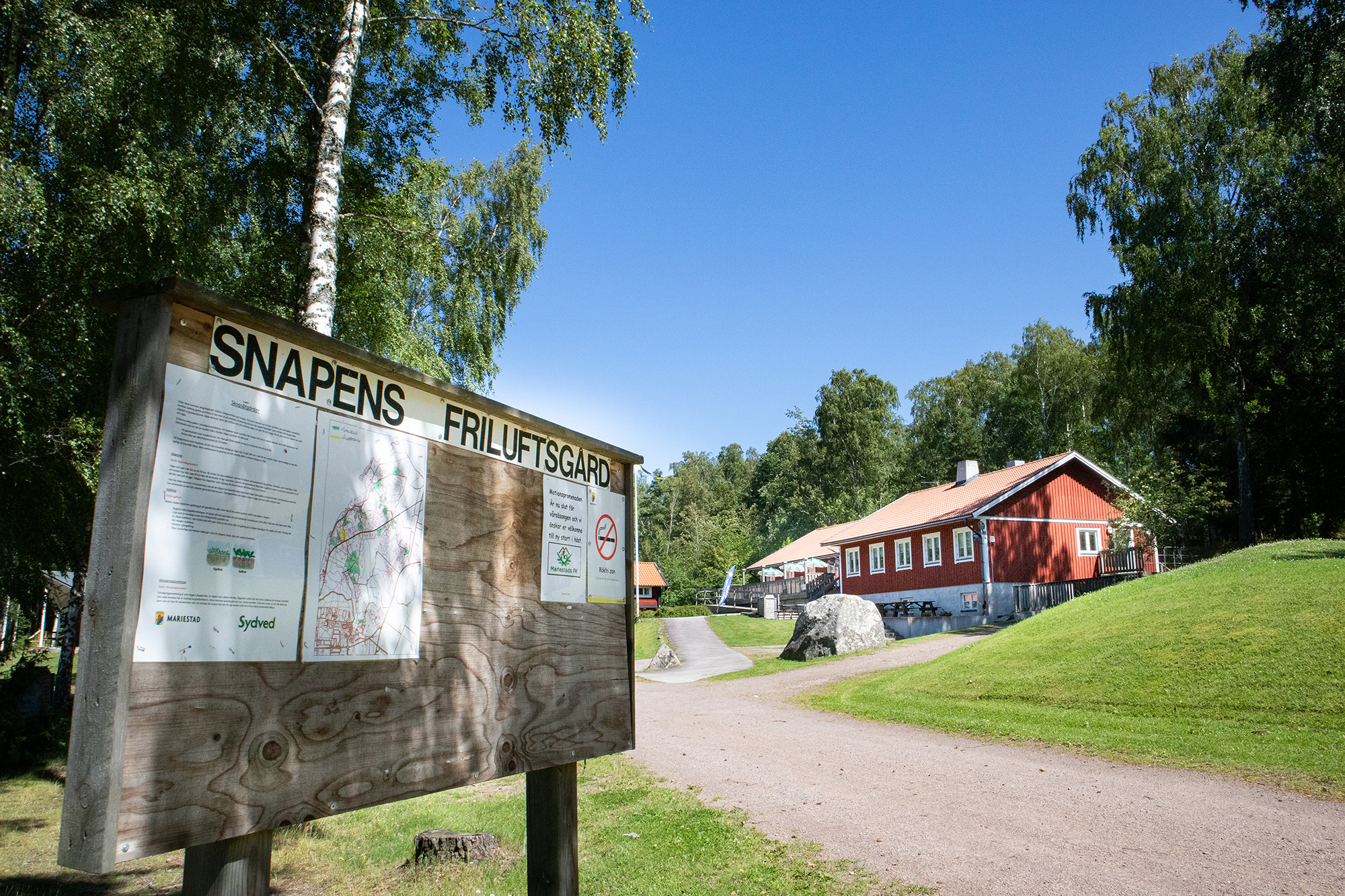 Anslagstavla med rubriken Snapens Friluftsgård i förgrunden. I bakgrunden syns en röd byggnad.